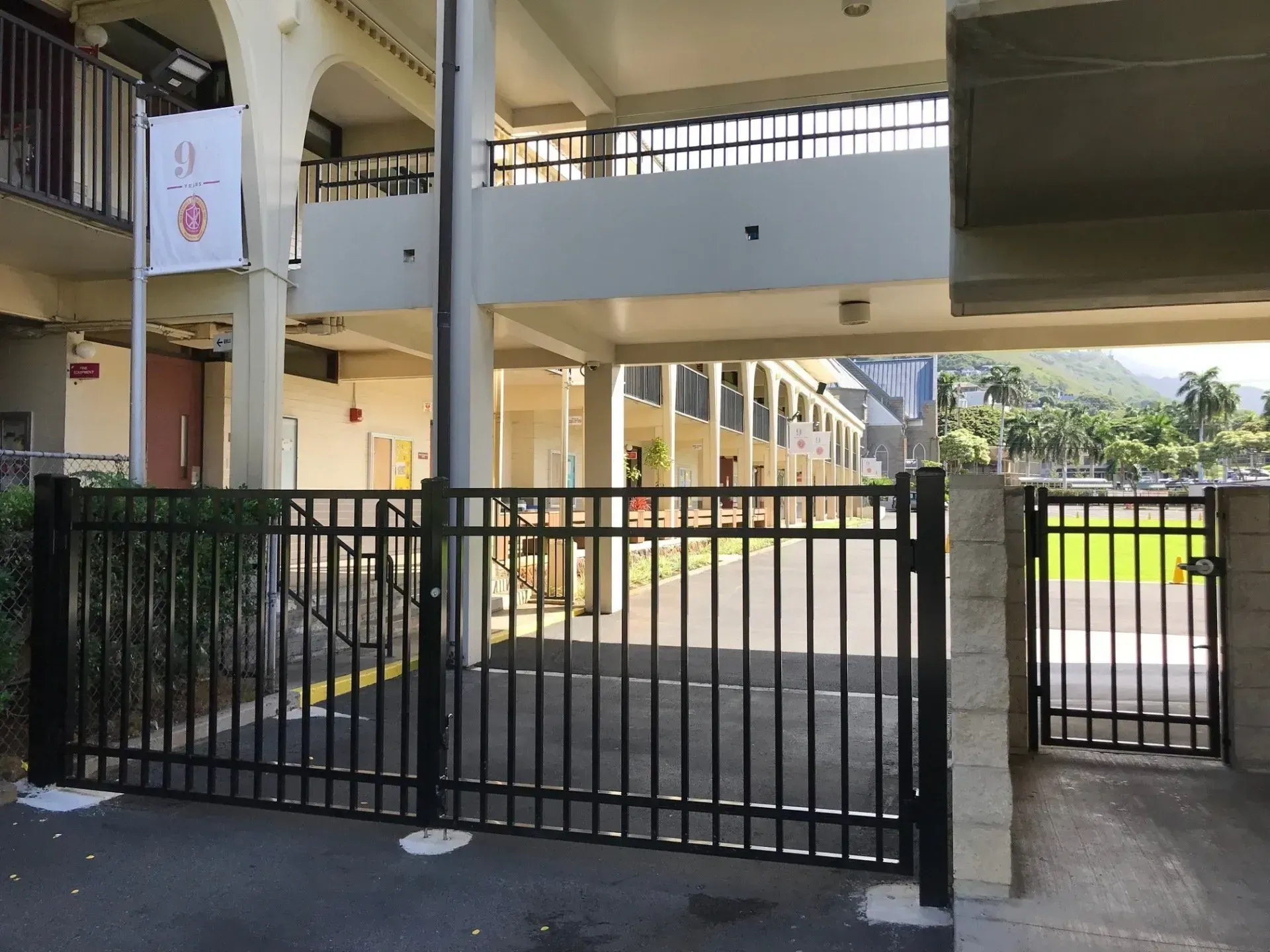 Black metal gate in front of a building with a white upper level and a glimpse of a green landscape.