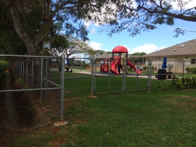 A fenced playground with a red slide and green grass. Houses and trees are in the background.