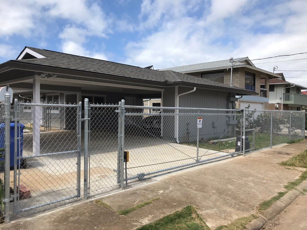 Chain-link fence with gate in front of a house with a carport and driveway under a cloudy sky.