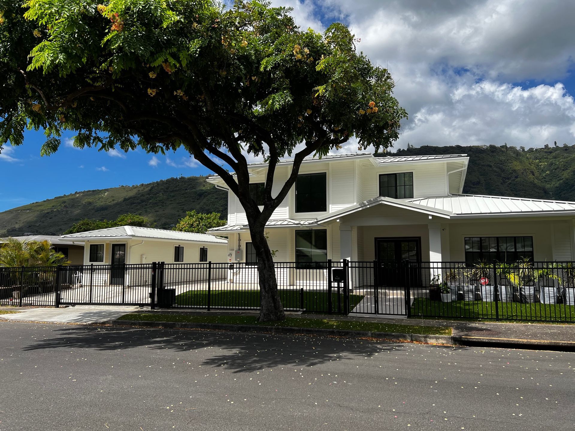 Two-story white house with a smaller adjacent structure, black fence, and tree. Mountains in the background.