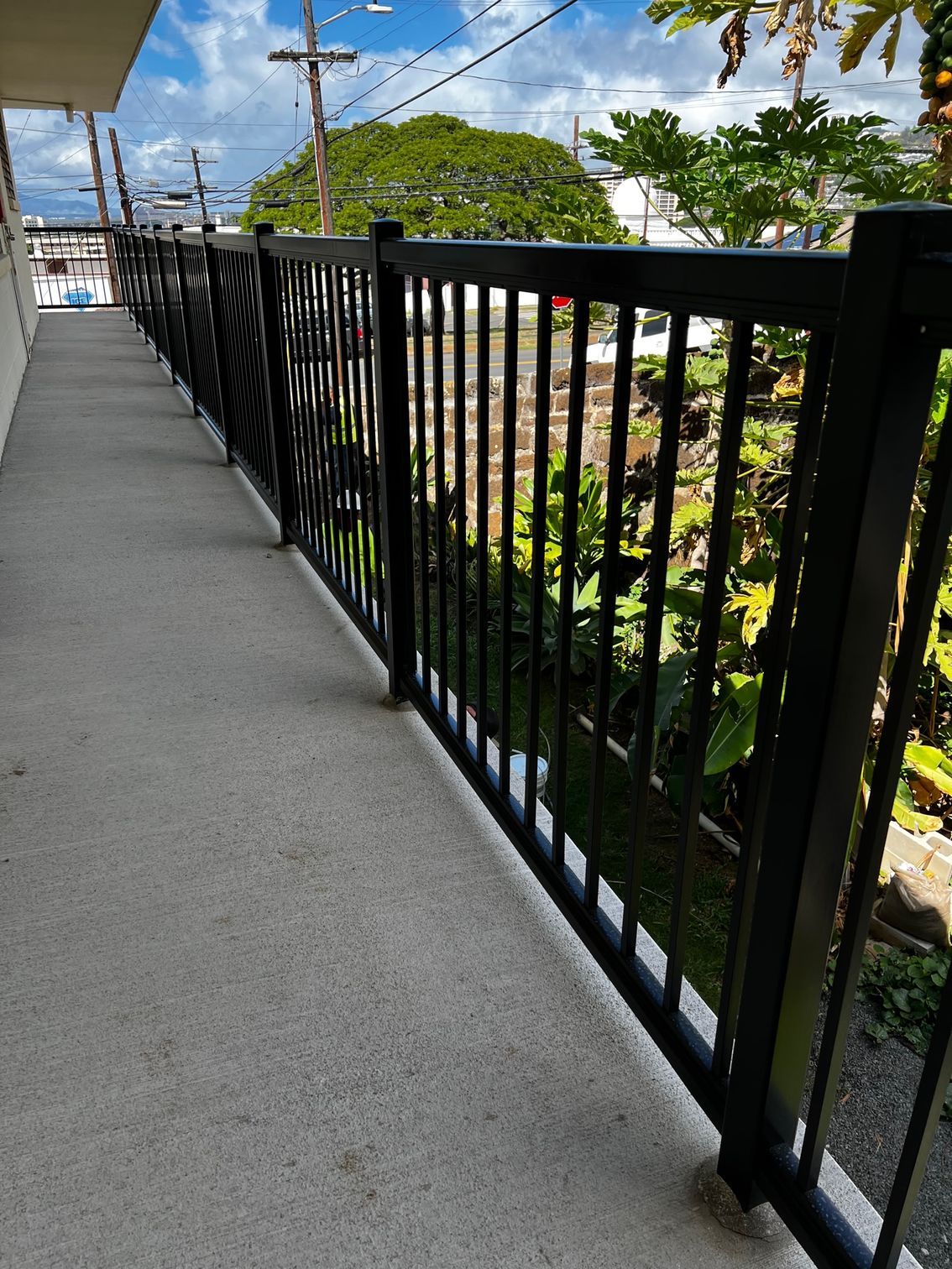 Black metal railing along a concrete walkway, with a green foliage backdrop and blue sky.