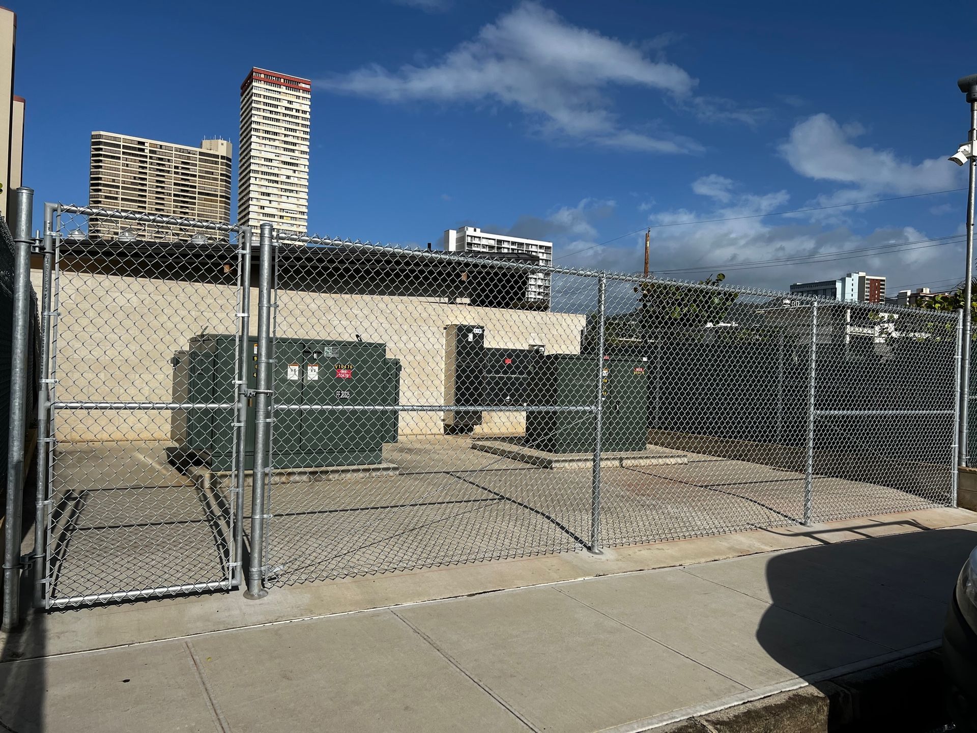 Chain-link fenced electrical equipment enclosure on a concrete pad; background buildings under a partly cloudy sky.