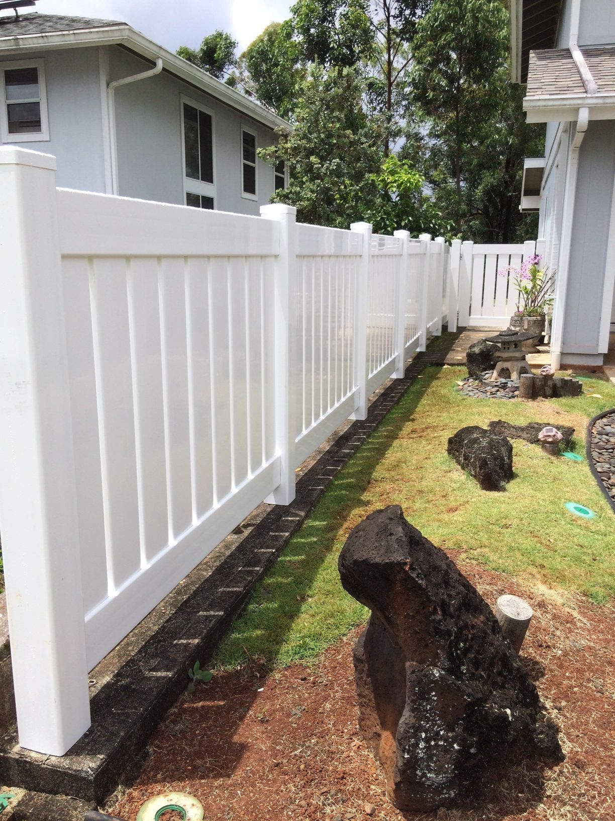 White vinyl fence bordering a yard with grass, brown soil, and large black rocks.