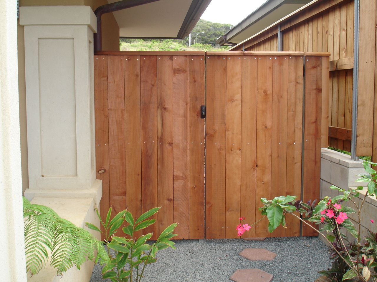 Wooden gate, tan and brown, between a pillar and fence, with plants and gravel below.