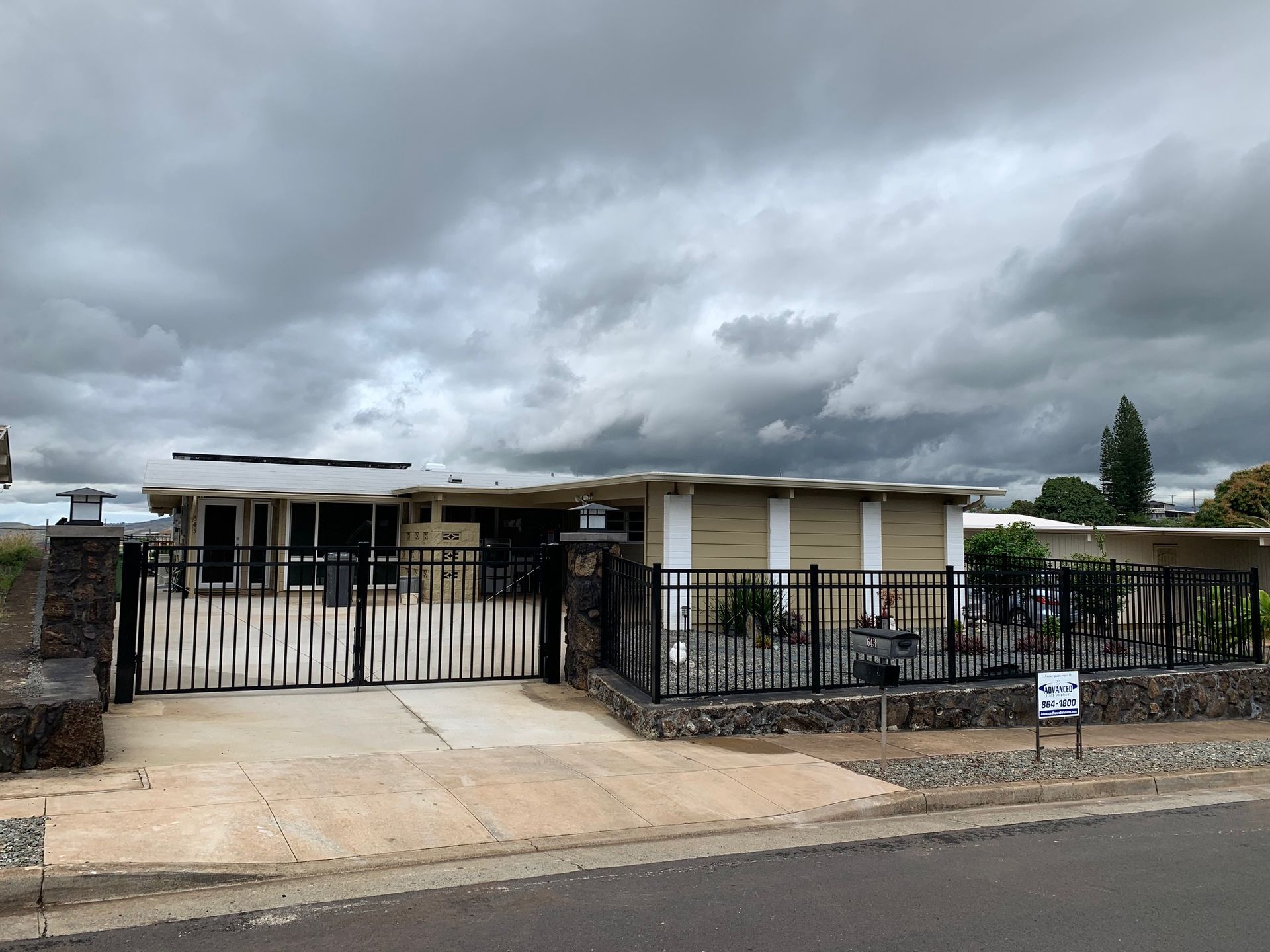 A one-story beige house behind a black metal fence with a gate. Gray, cloudy sky.