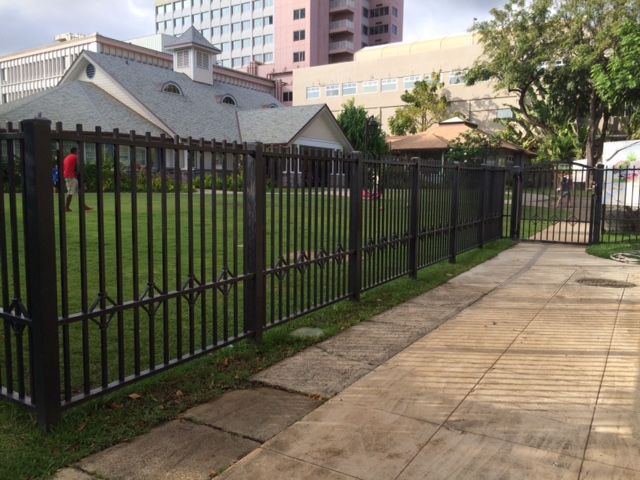 Black metal fence bordering a green lawn with a pathway. Buildings in background.