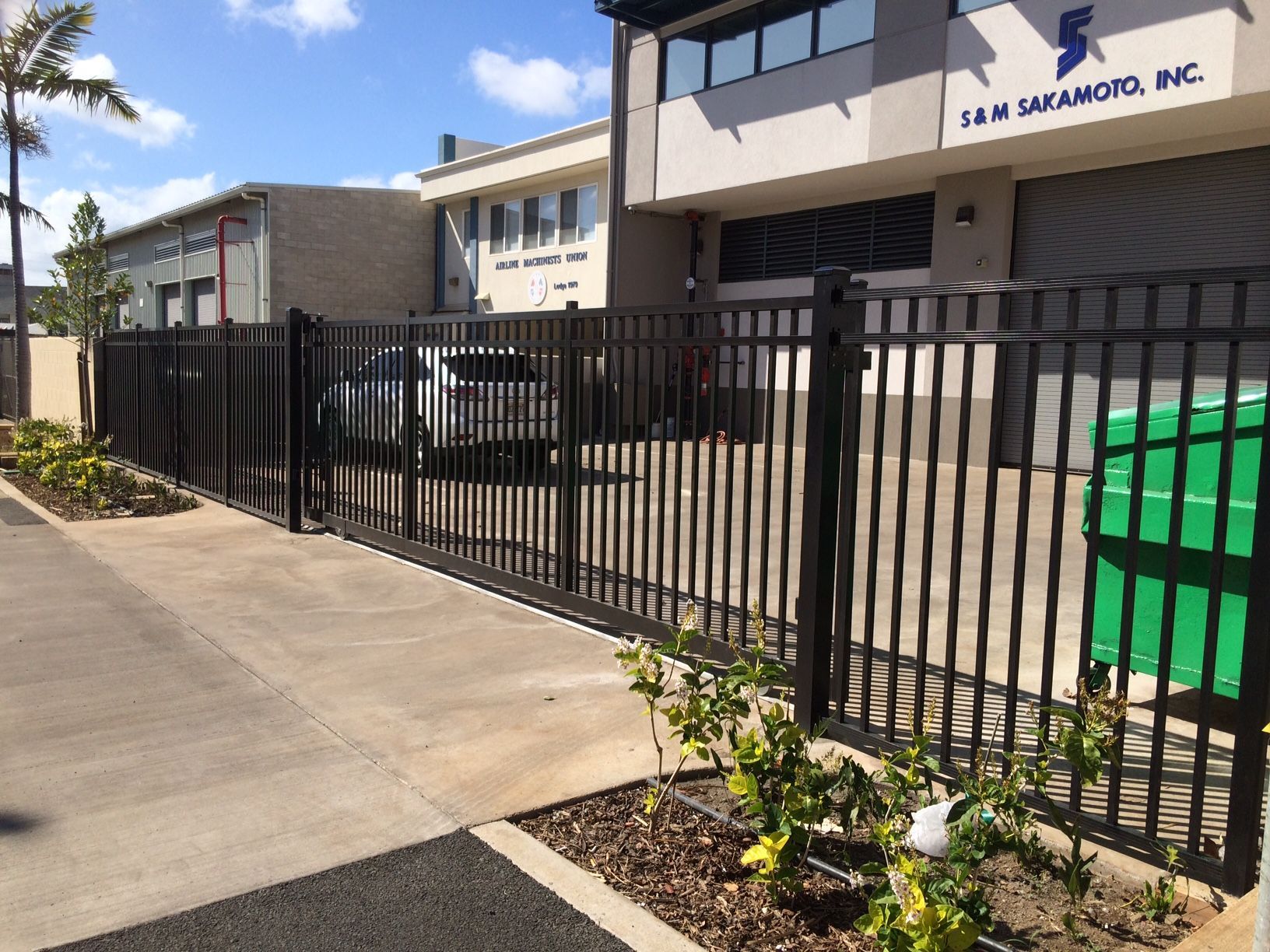 Black metal fence in front of a building with a driveway and plants.