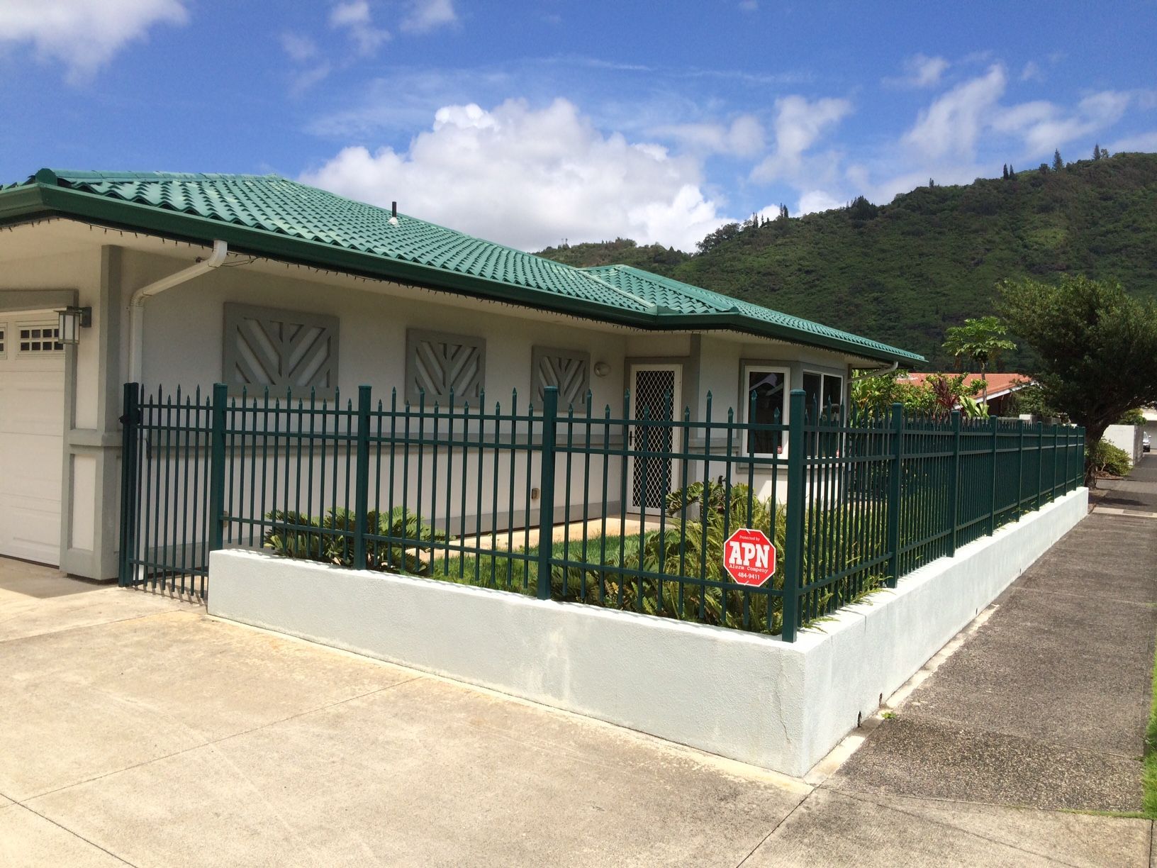 Light gray house with teal roof, green metal fence, driveway, sidewalk, and mountain backdrop.