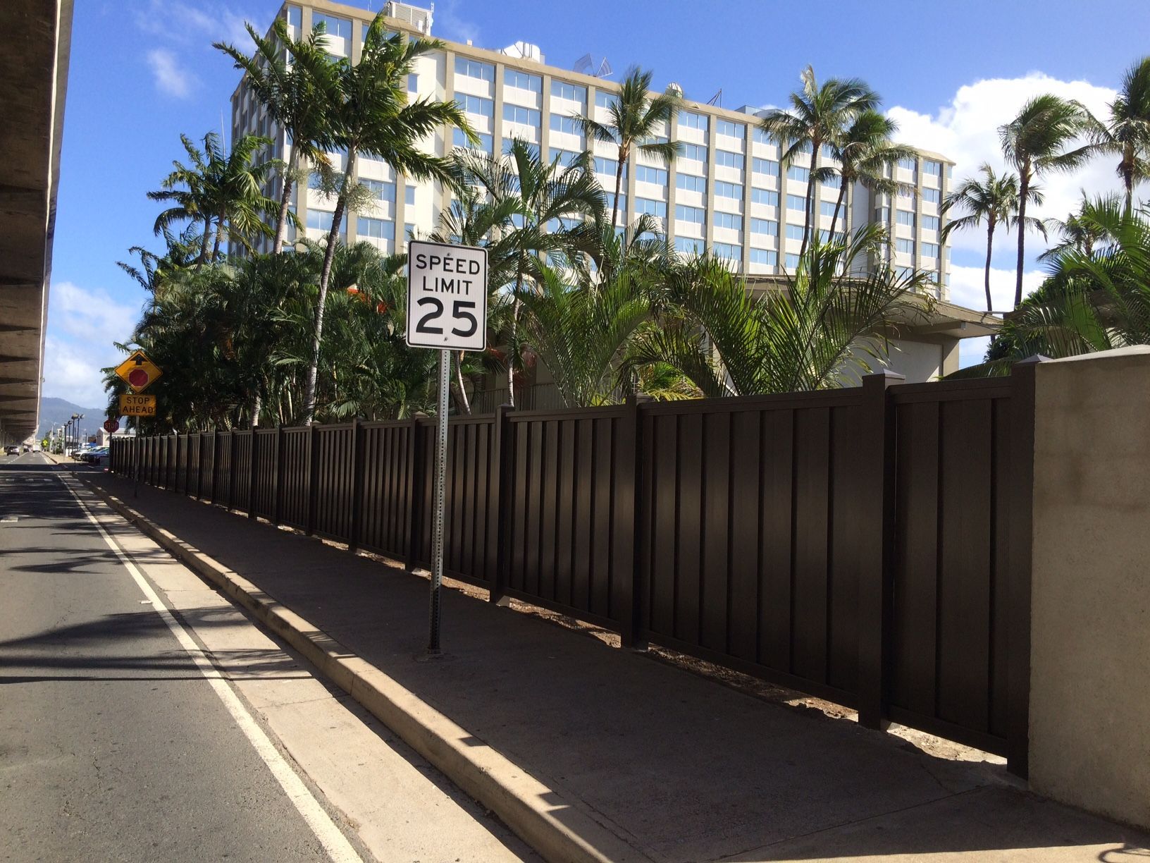 Street scene with a speed limit sign, brown fence, sidewalk, palm trees, and a tall building.