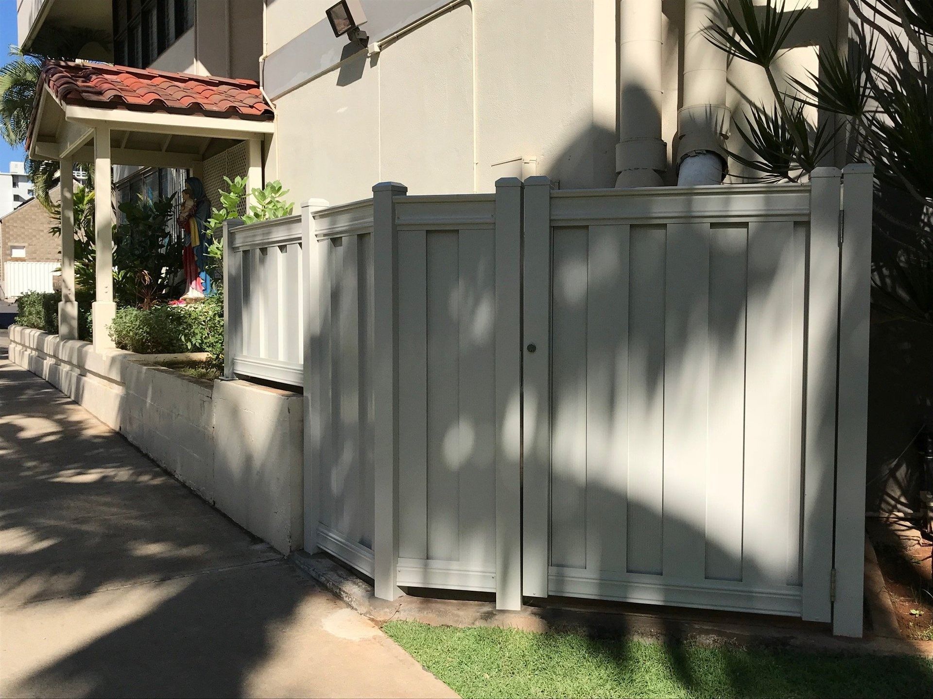 Beige fence next to a building on a sidewalk. Sun casts shadows.