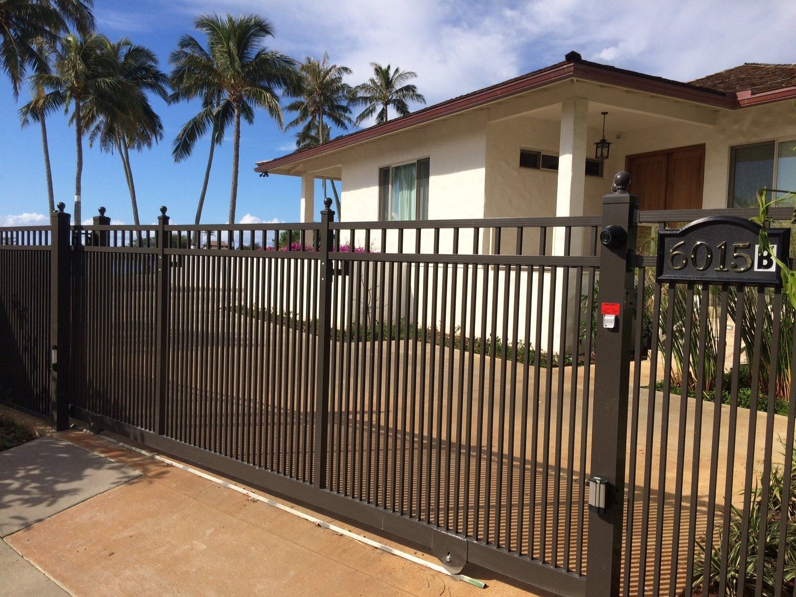 Brown metal gate in front of a white house with palm trees in the background. Address marker visible.