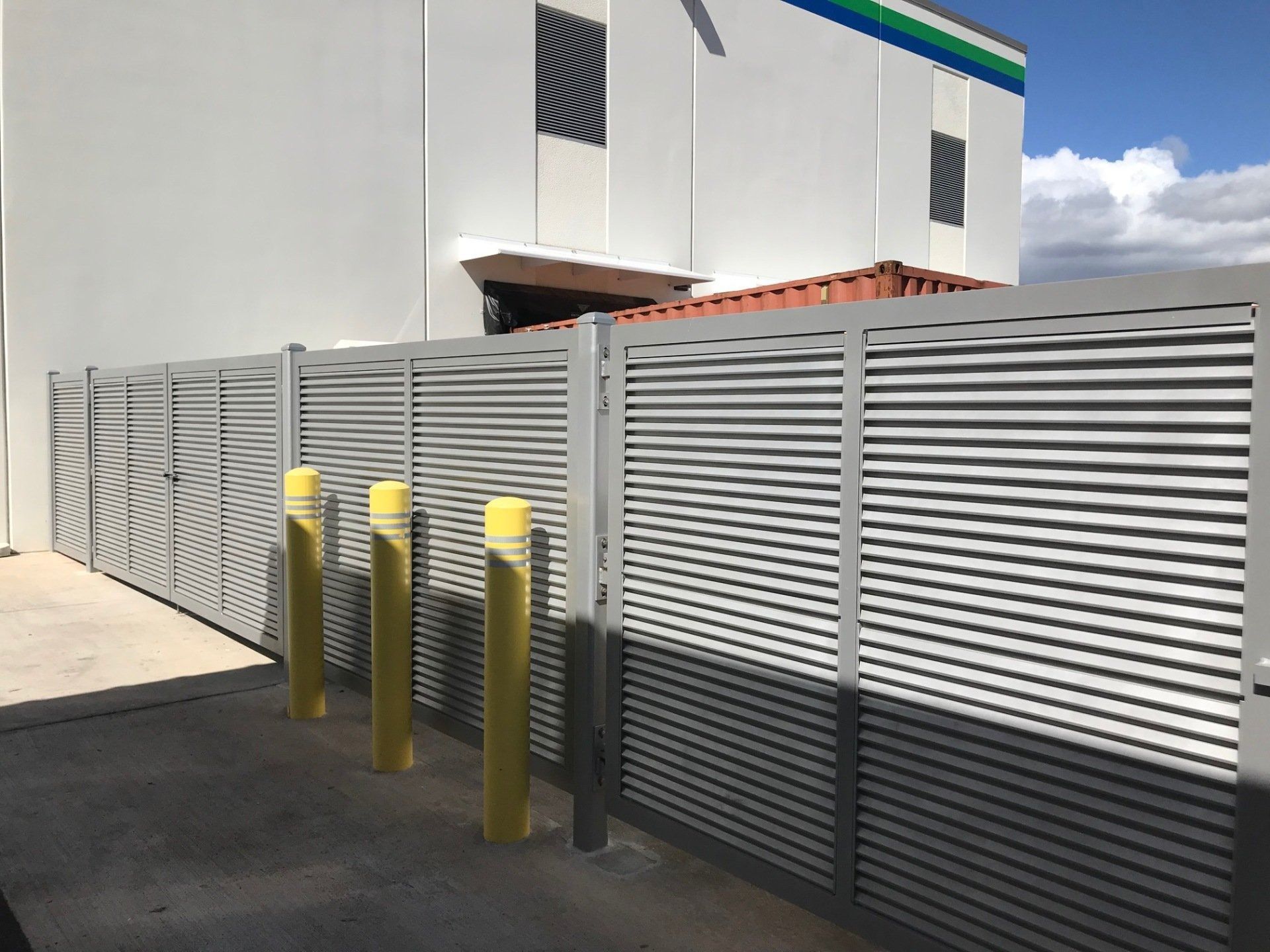Gray slatted metal fence with yellow bollards in front of a white building.