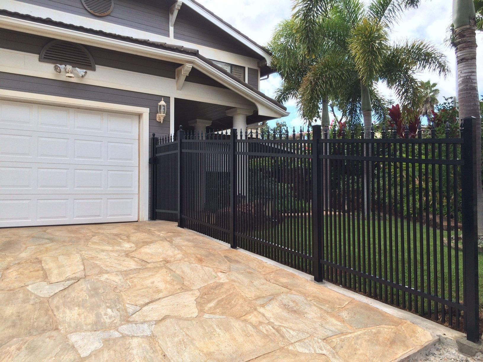 Black metal fence in front of a house, spanning across a driveway with a light brown, patterned surface.
