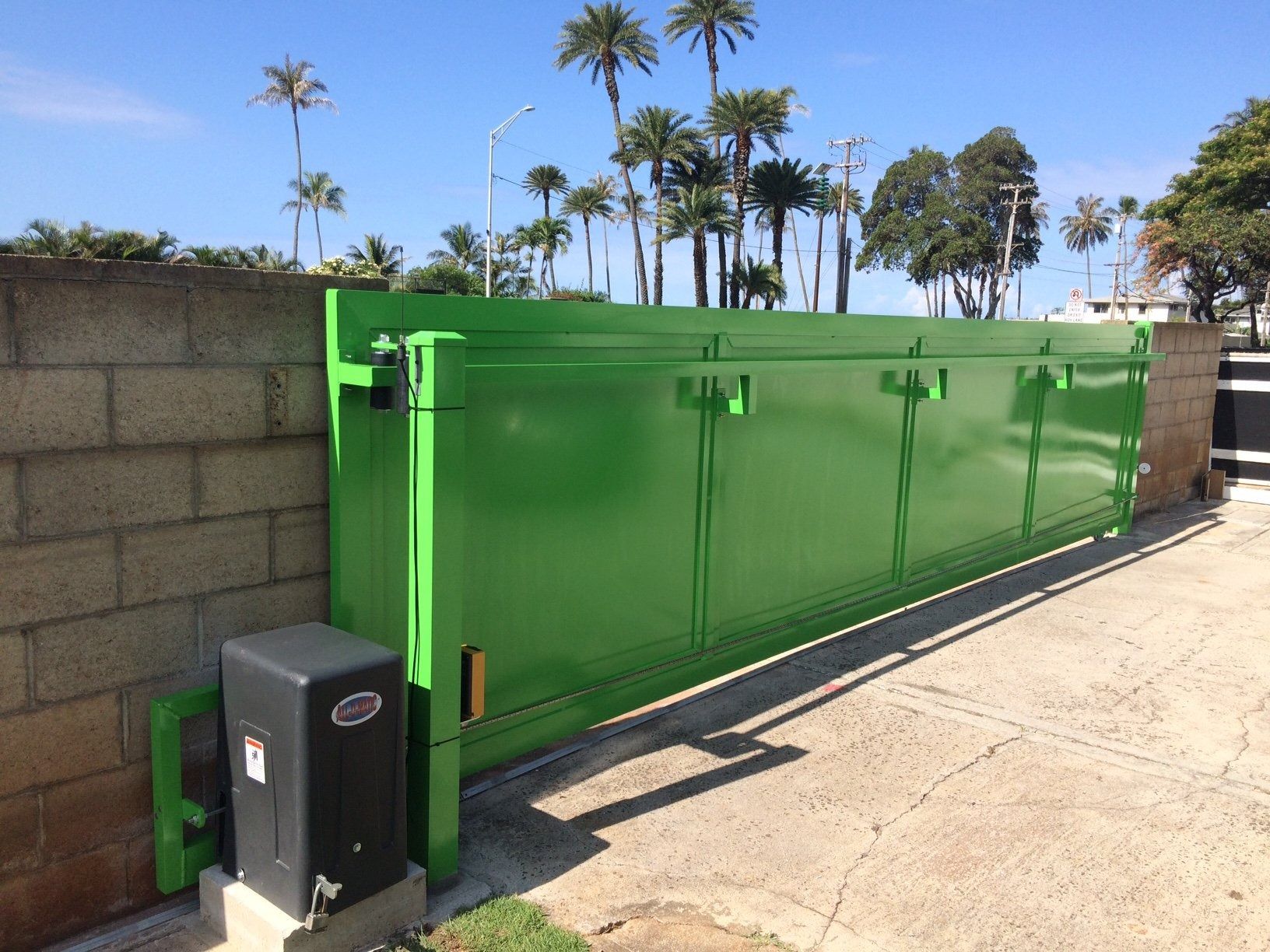 Green sliding gate next to a brick wall with an automatic opener. Palm trees in the background.