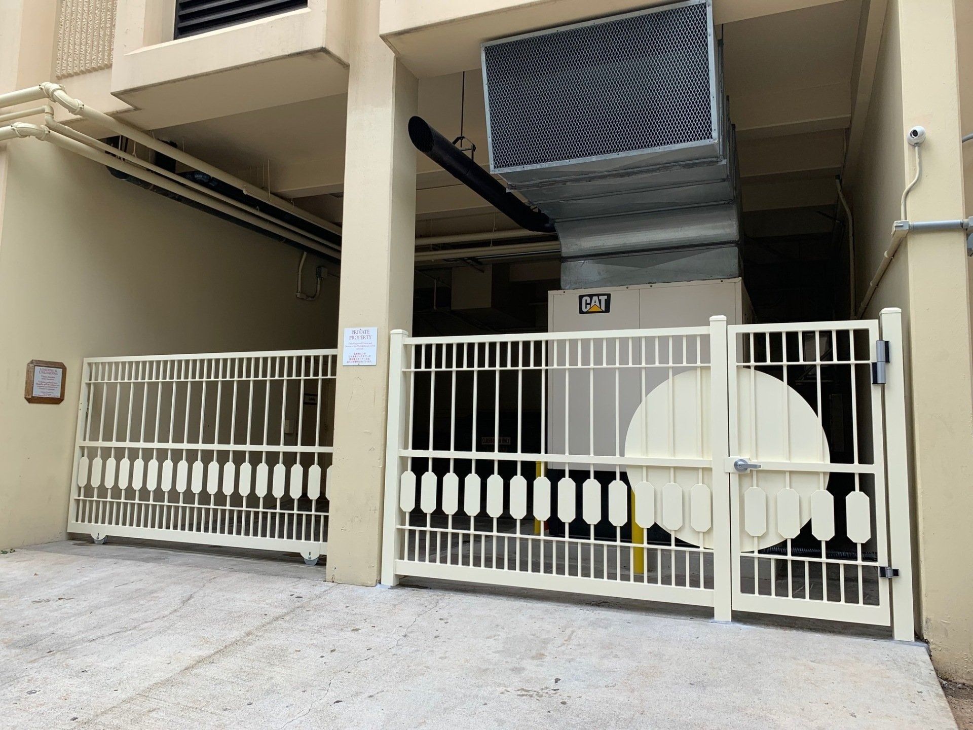 Cream-colored metal gates enclose a machinery area beneath a building. A large HVAC unit is visible.