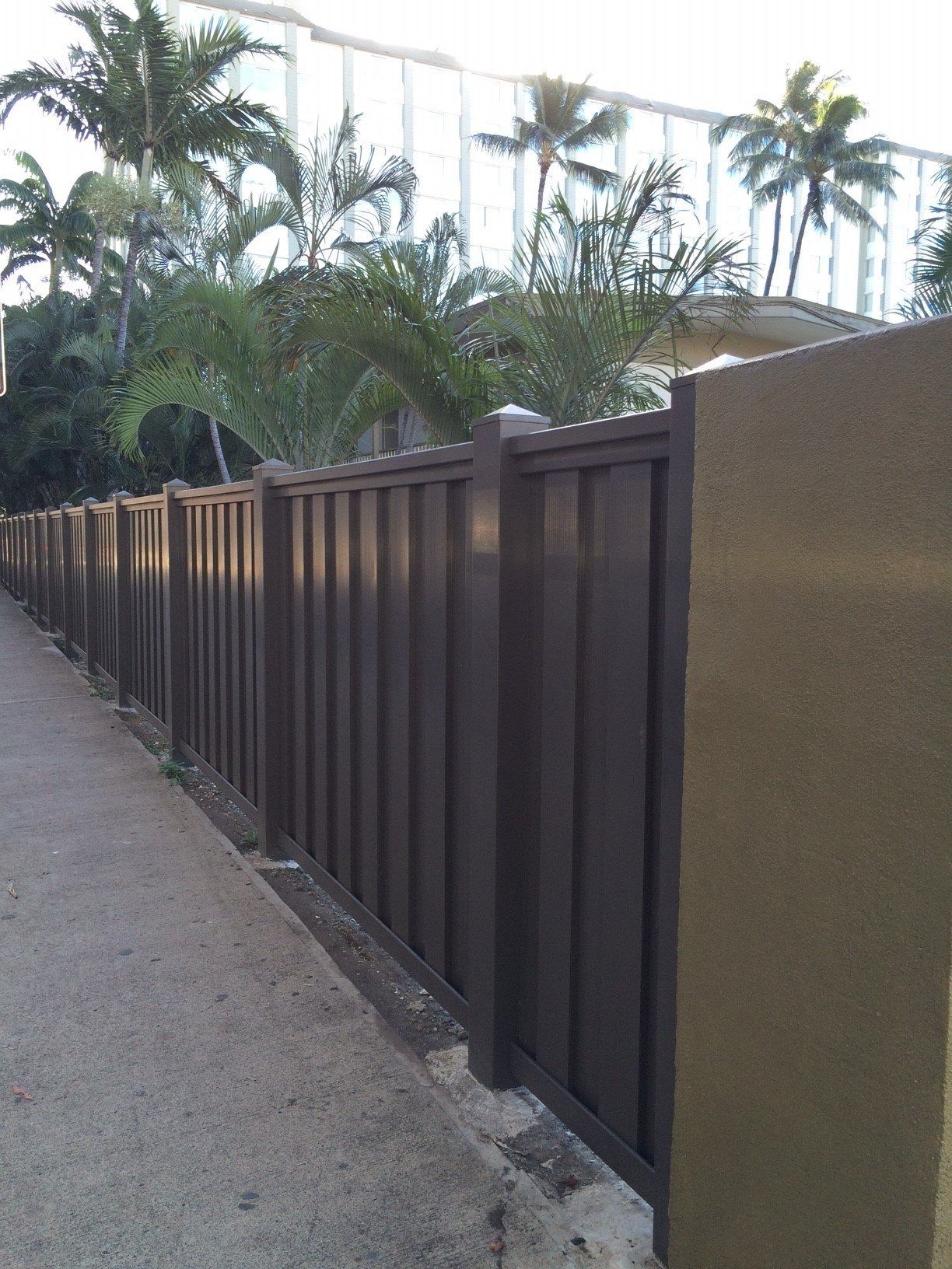 Brown fence along a sidewalk, with palm trees and a building in the background.