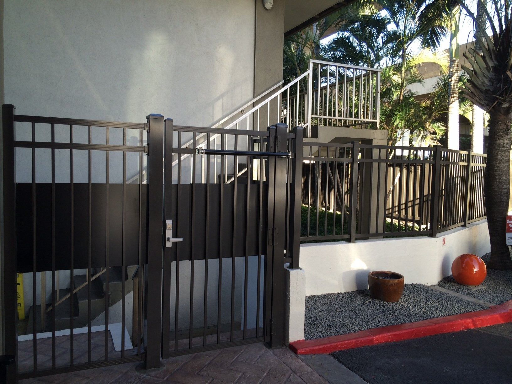 Black metal gate at building entrance with stairs. Brown ground, red curb, and palm tree.