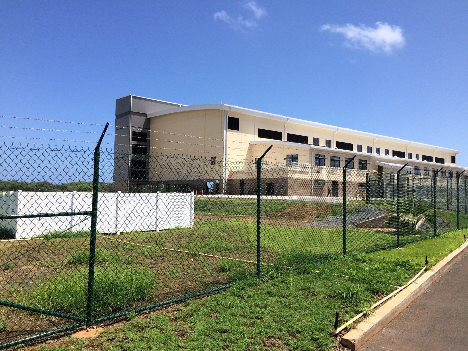 A light-colored building with a dark top is surrounded by a chain-link fence on a sunny day.