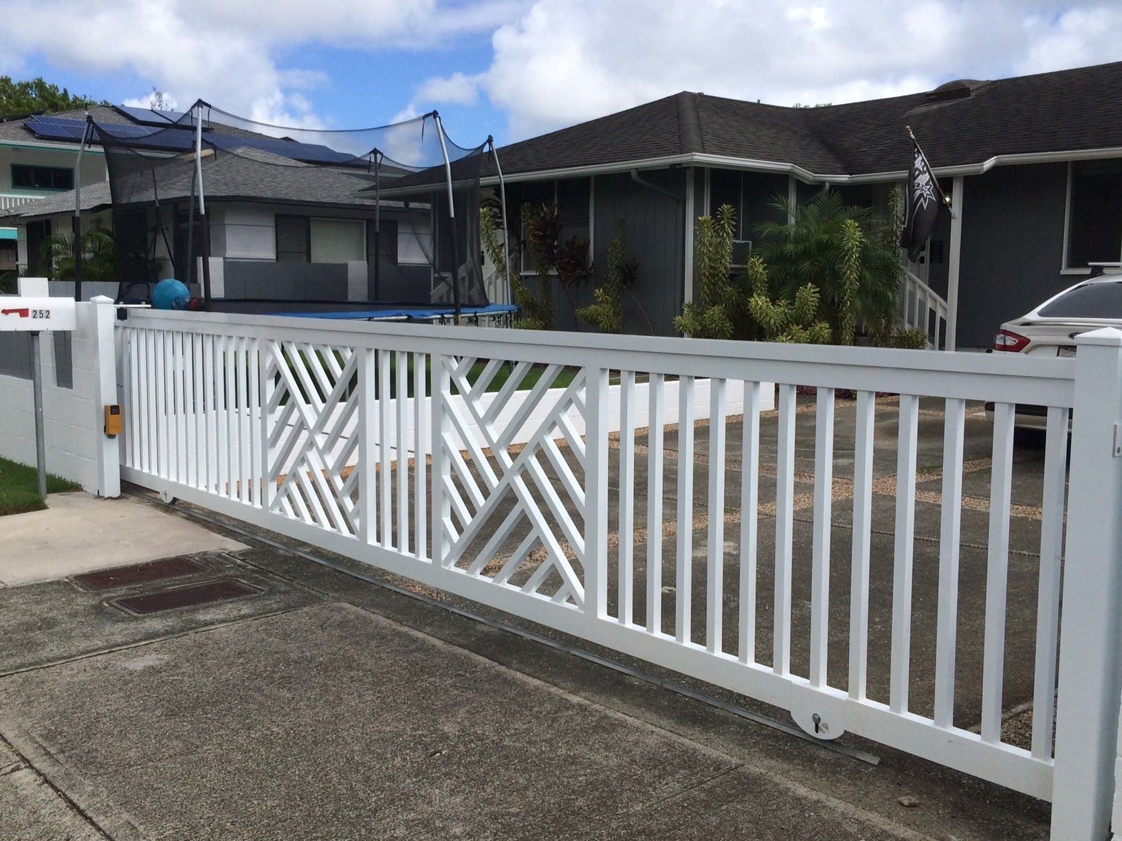 White decorative gate in front of a gray house and trampoline on a sunny day.