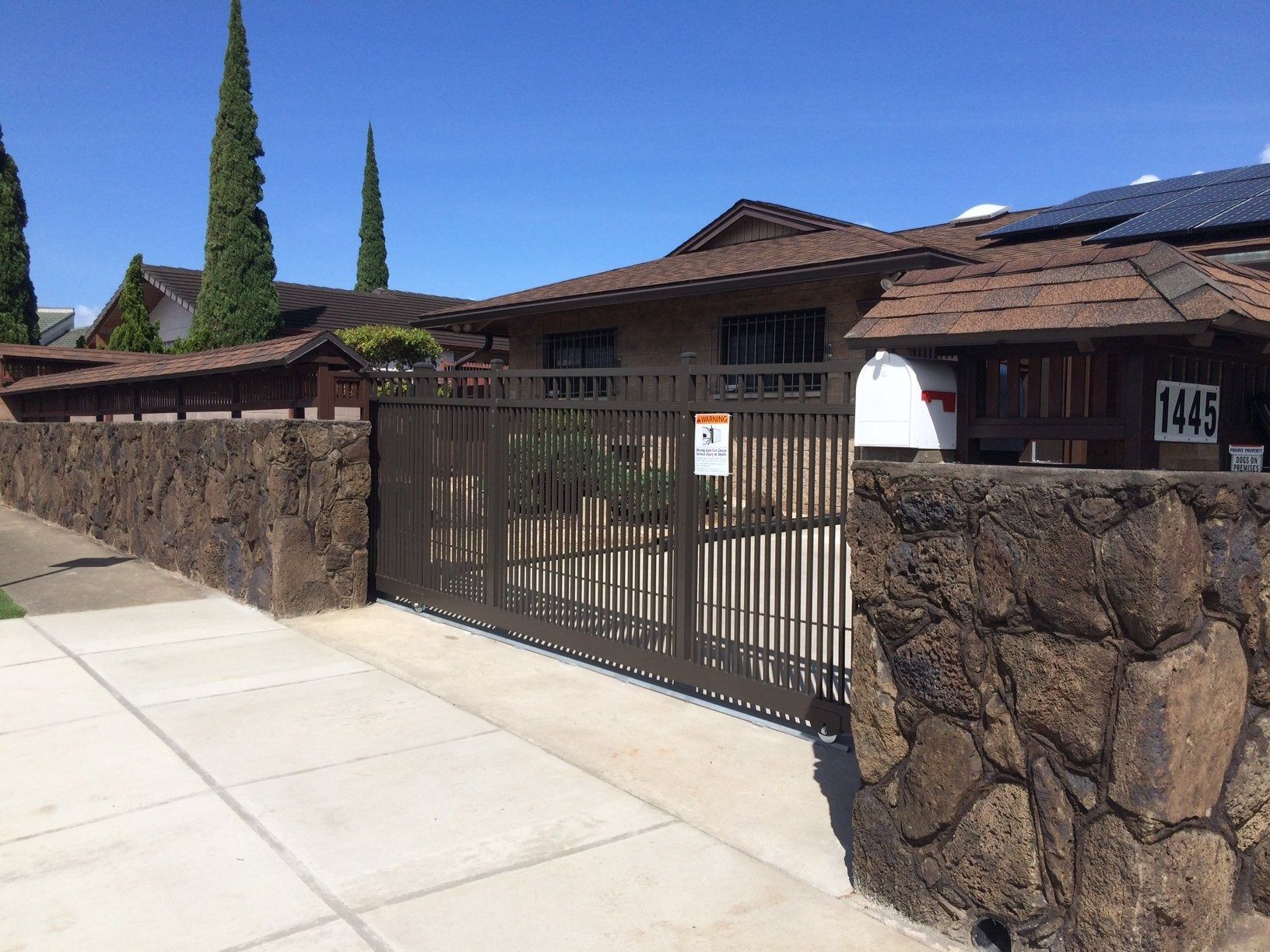 A brown gate and stone wall enclose a house with a brown roof and solar panels.