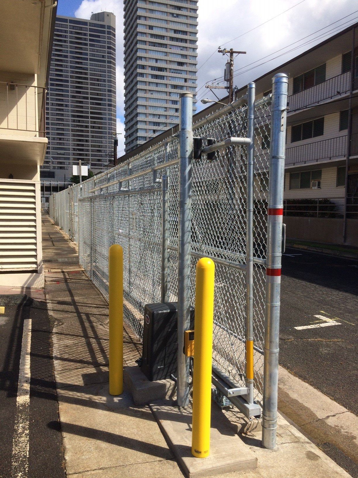 Chain-link fence with yellow bollards. Tall buildings in the background. Asphalt pavement.