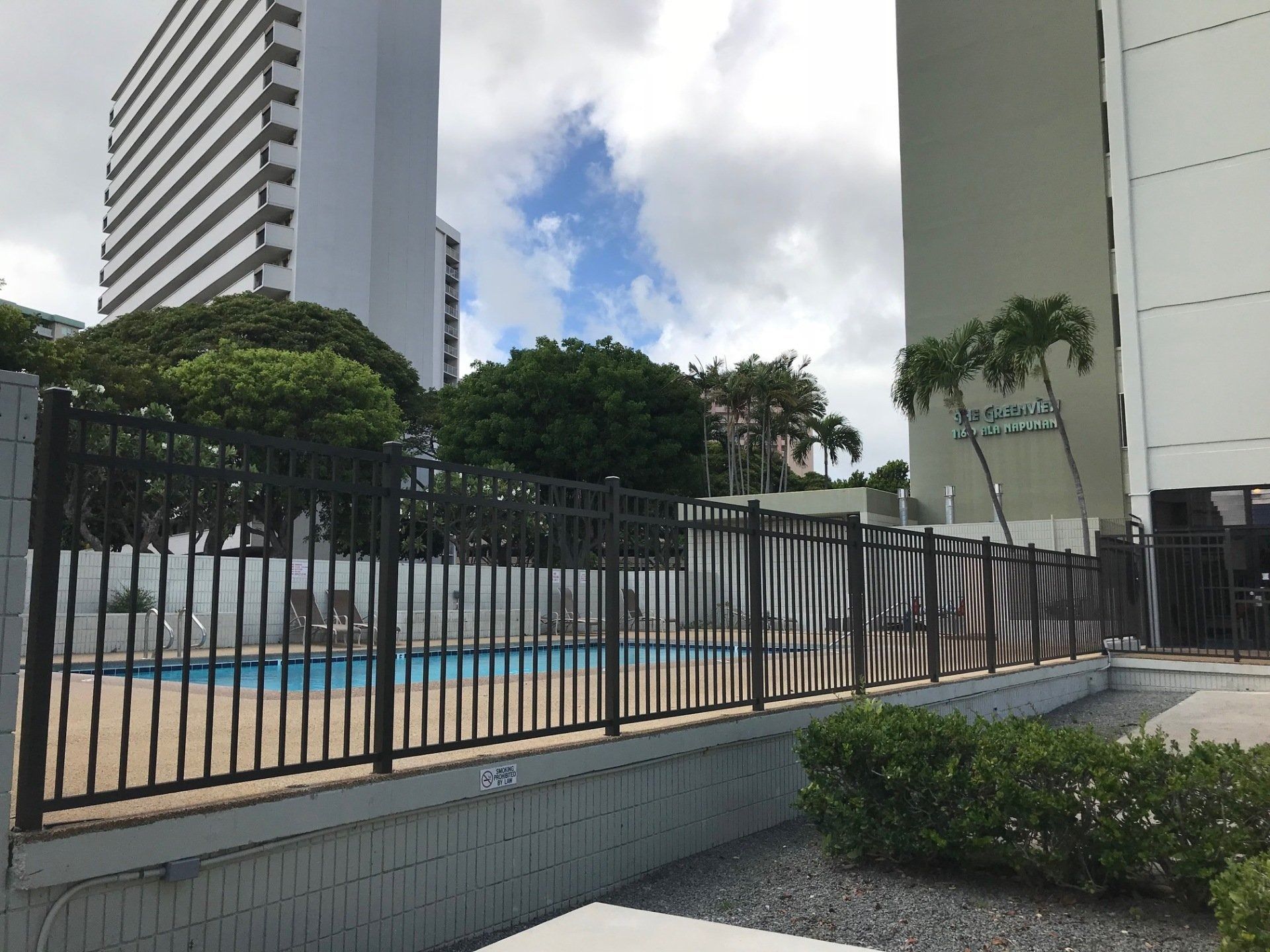 Pool behind a black metal fence, flanked by buildings, under a cloudy sky.