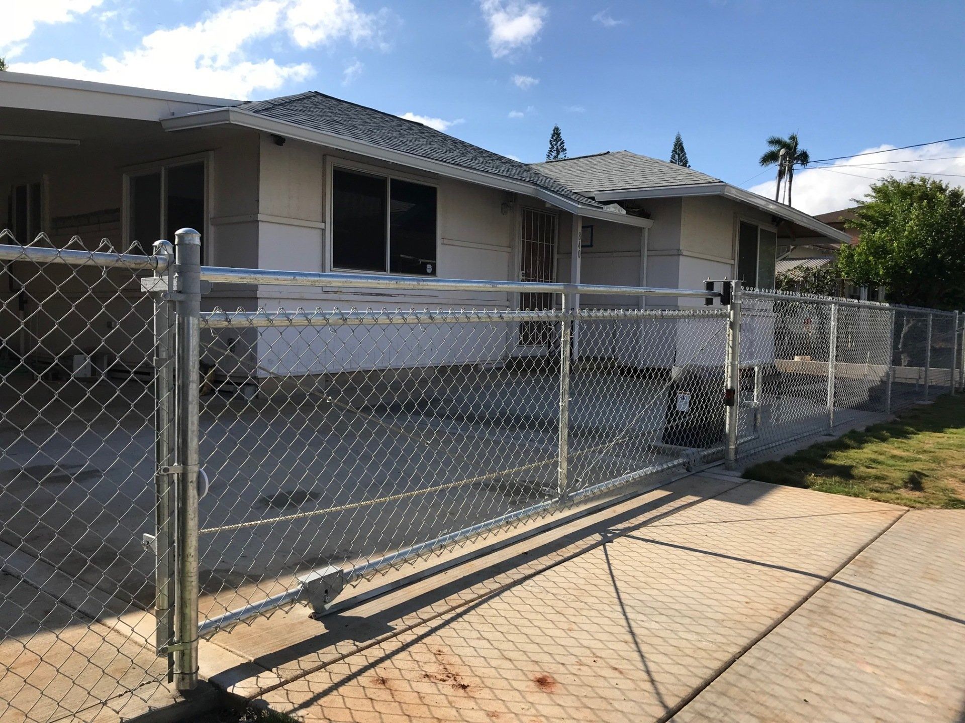 A residential home with a chain-link fence in front. Gray roof, beige siding, sidewalk. Blue sky.