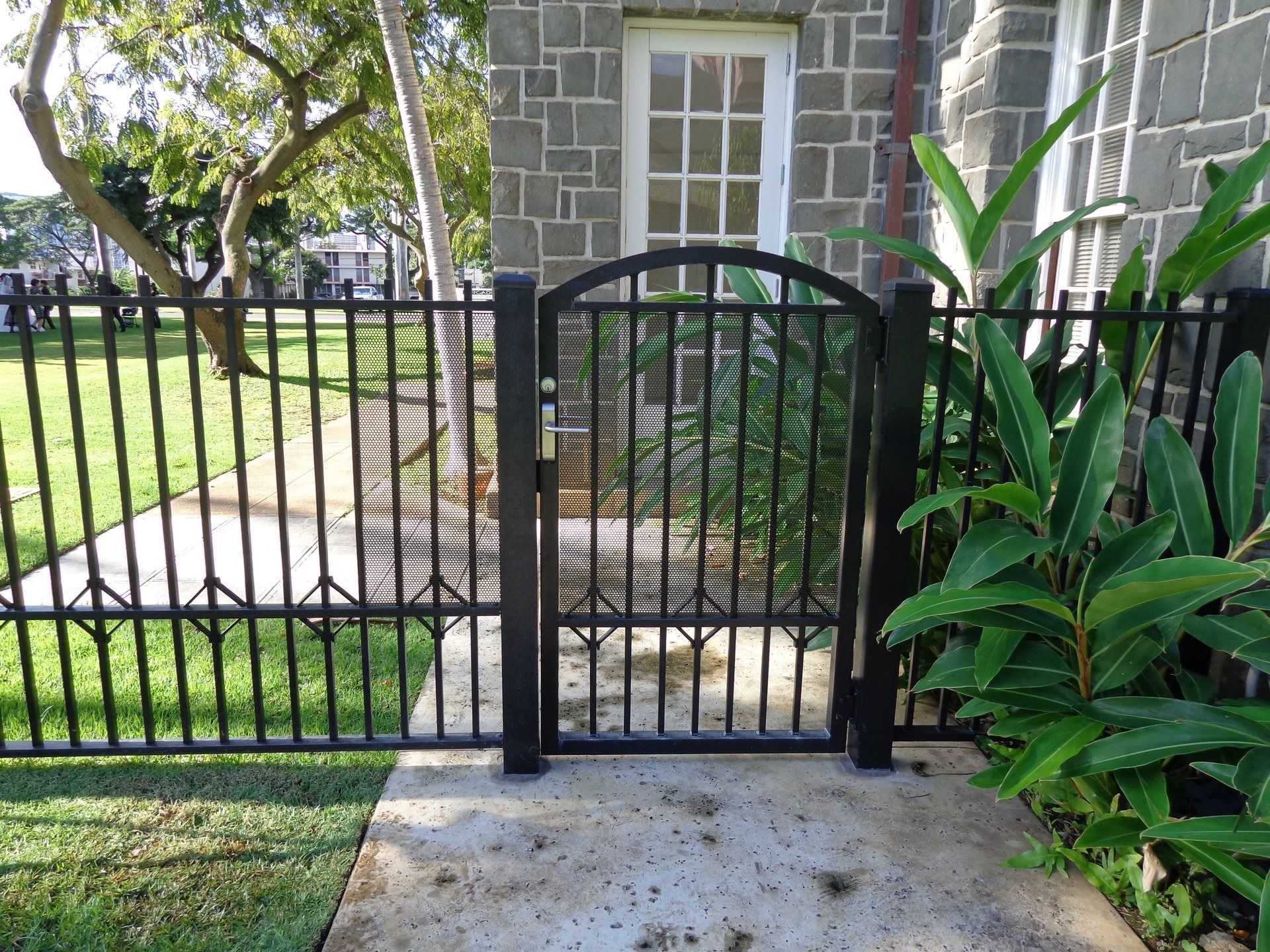 Black metal gate in a matching fence with a stone building in the background and greenery on the side.