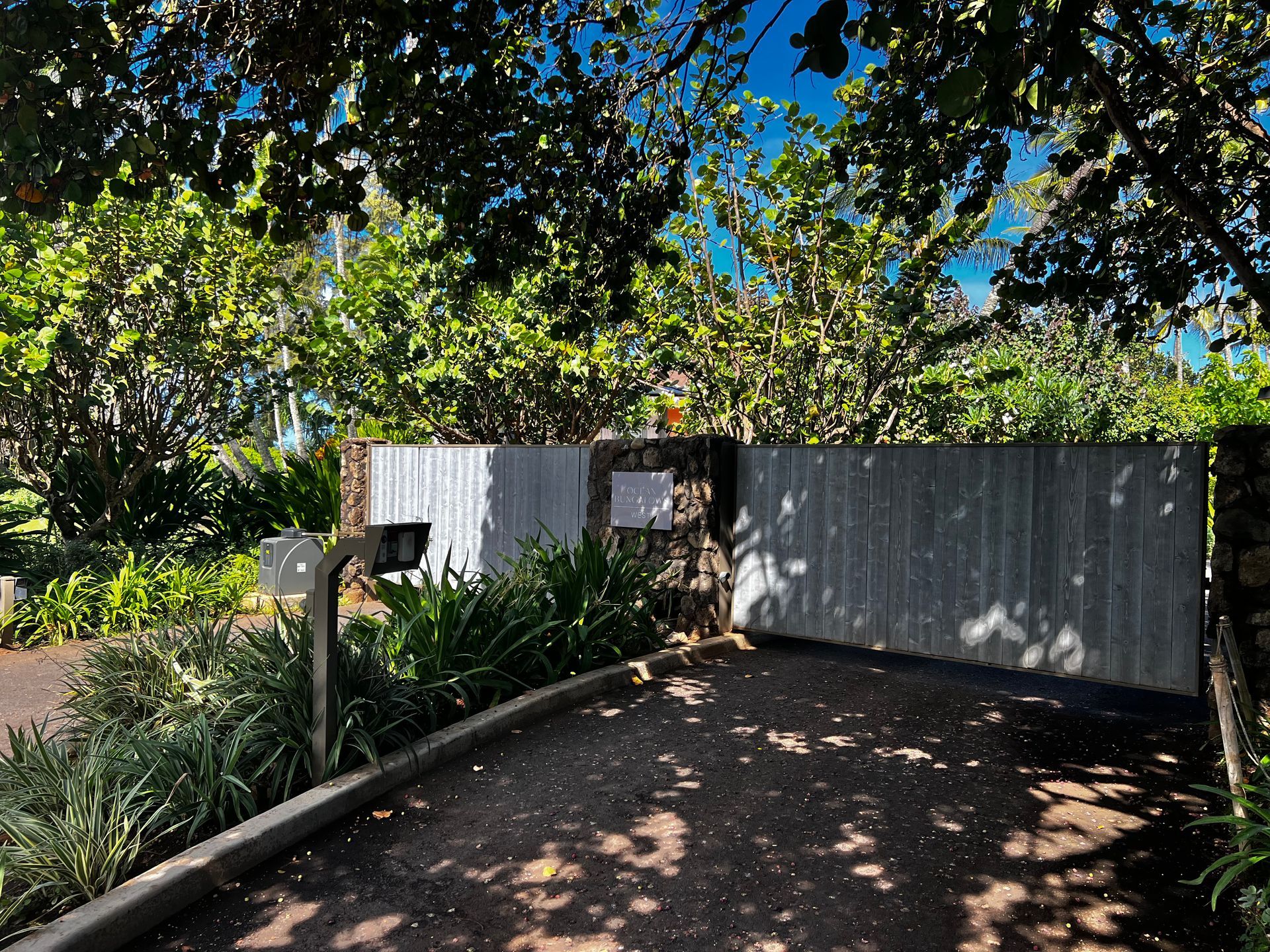 A driveway leads to a wooden gate, behind which is lush greenery, with a mailbox and sign post in front.