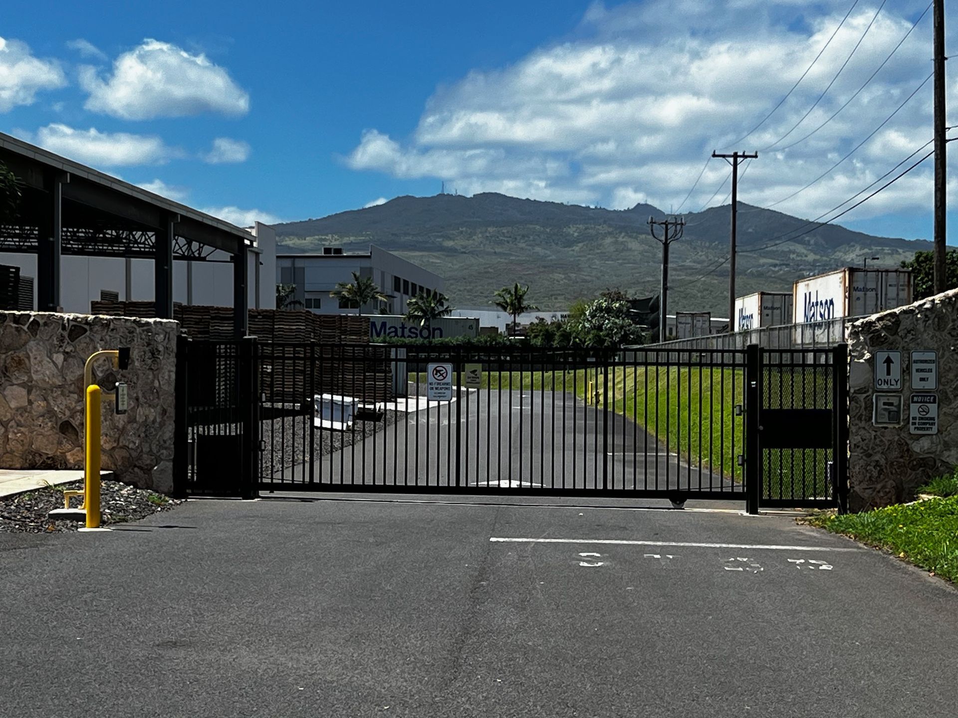Black gated entrance on a paved road, leading to buildings and mountain in the background, blue sky with clouds.