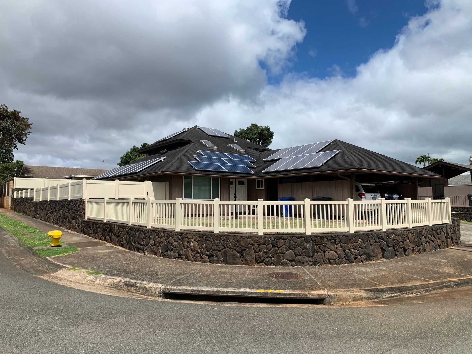House with solar panels on roof, surrounded by a fence atop a dark stone wall; cloudy sky.