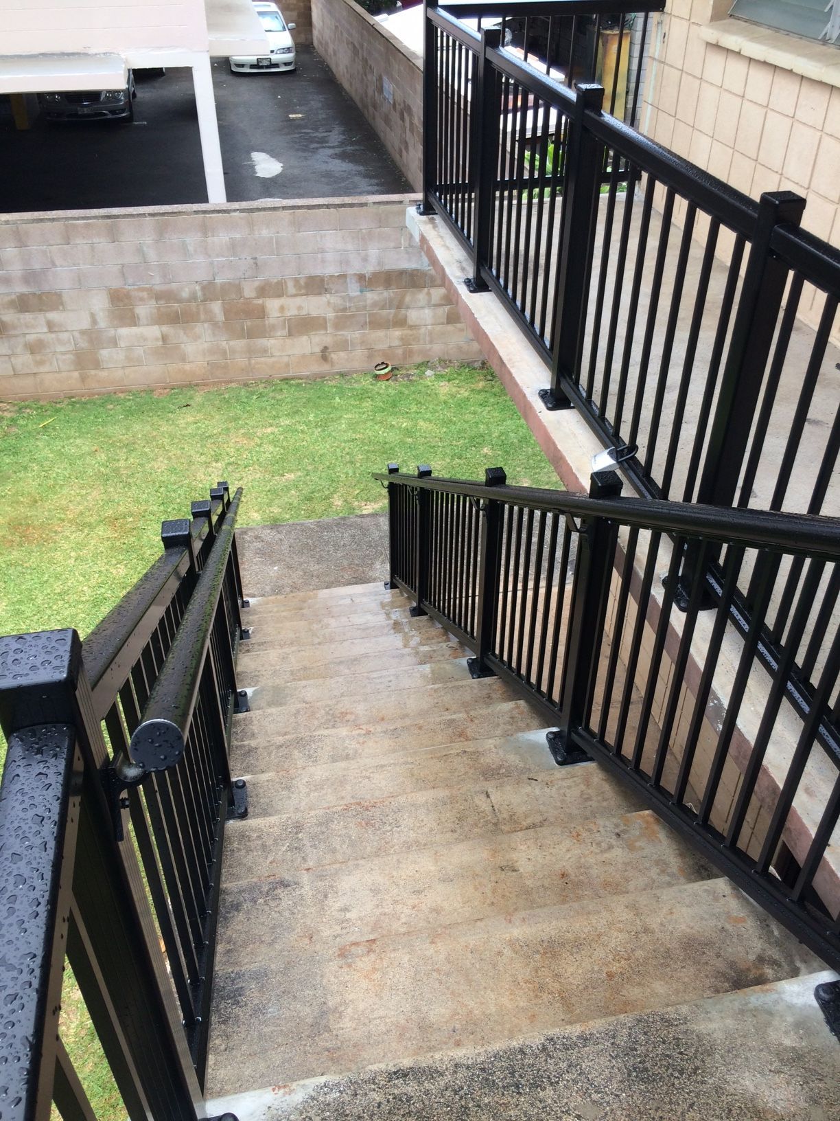 Concrete stairs with black railings descend into a grassy area, alongside a tan wall.