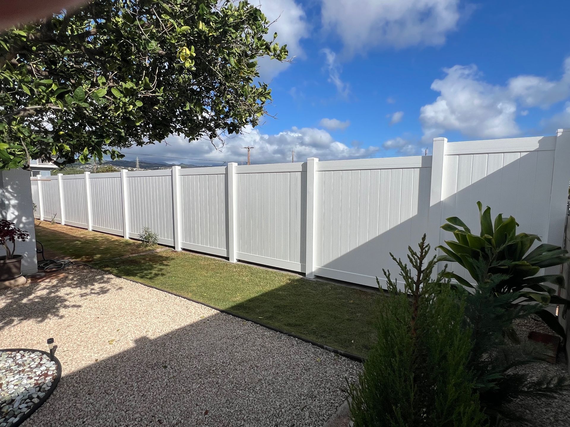 White vinyl fence along green grass and gravel. Blue sky with clouds.