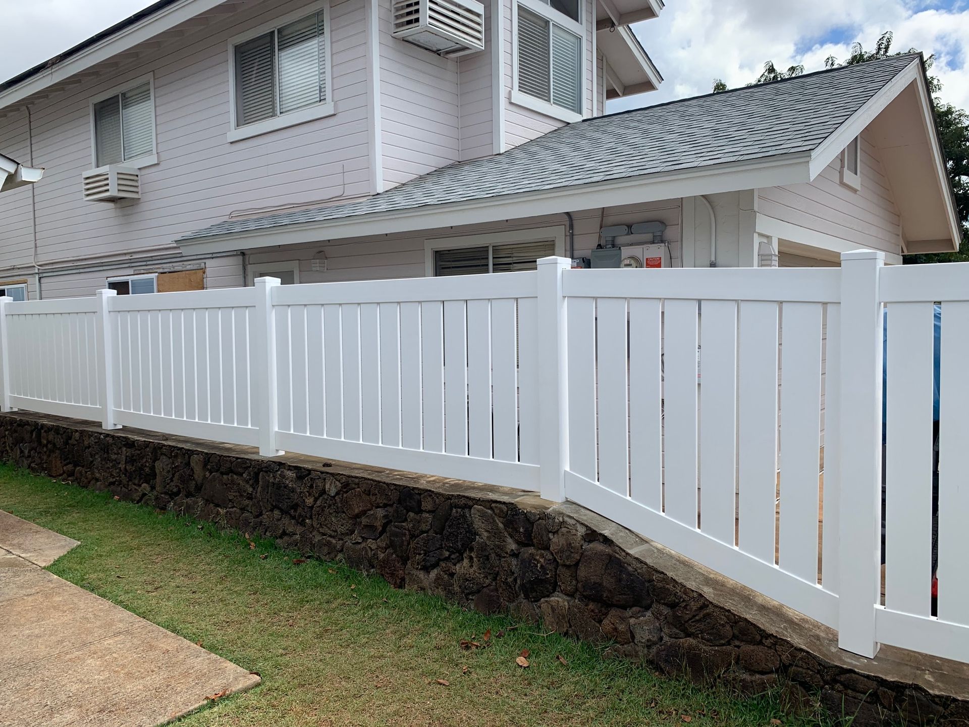White fence in front of a white house with a gray roof, set on a low stone wall and green lawn.