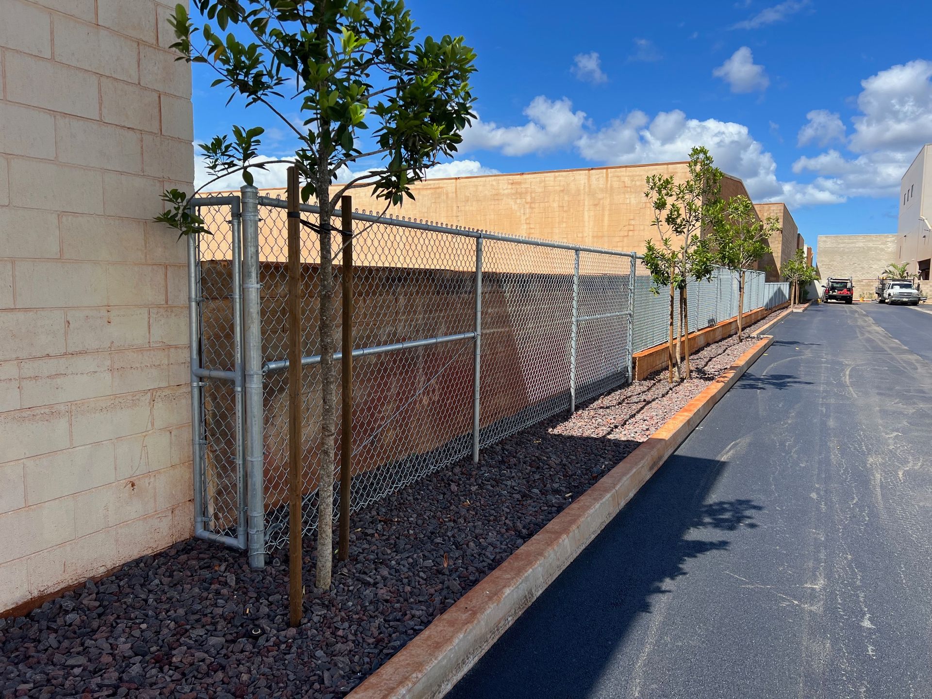 Chain link fence and trees along a paved driveway next to a tan building under a blue sky.