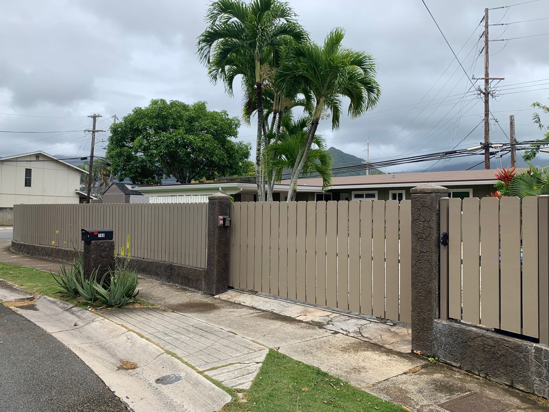 Beige wooden fence with concrete pillars in front of a house, palm trees in the background, cloudy sky.