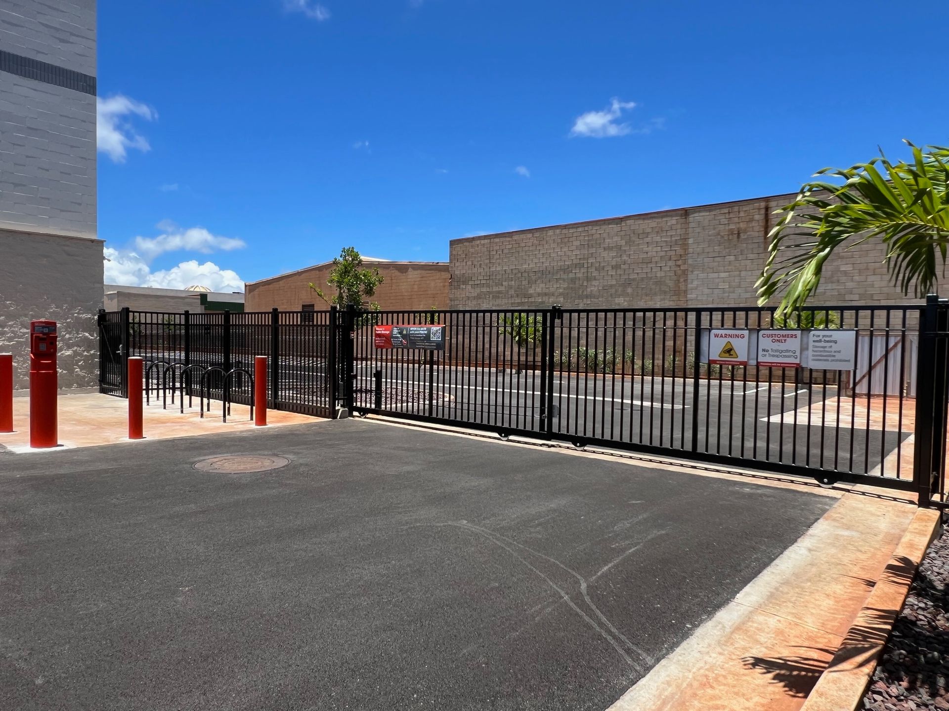 Black metal gate at entrance to a paved area, red bollards on left, brick buildings and blue sky in background.