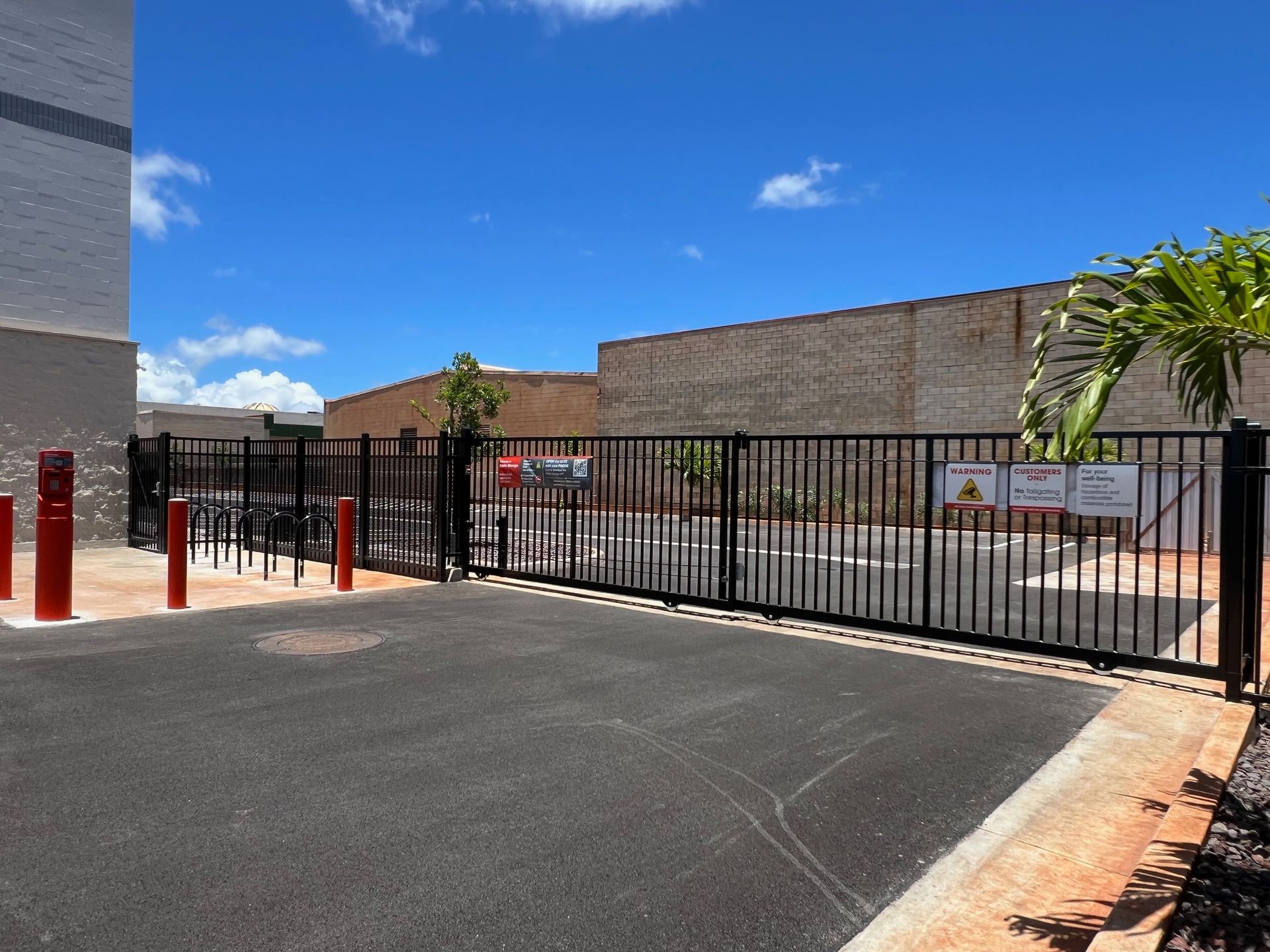 Black metal gate at the entrance to a paved area, red bollards on the left, brick building in background.