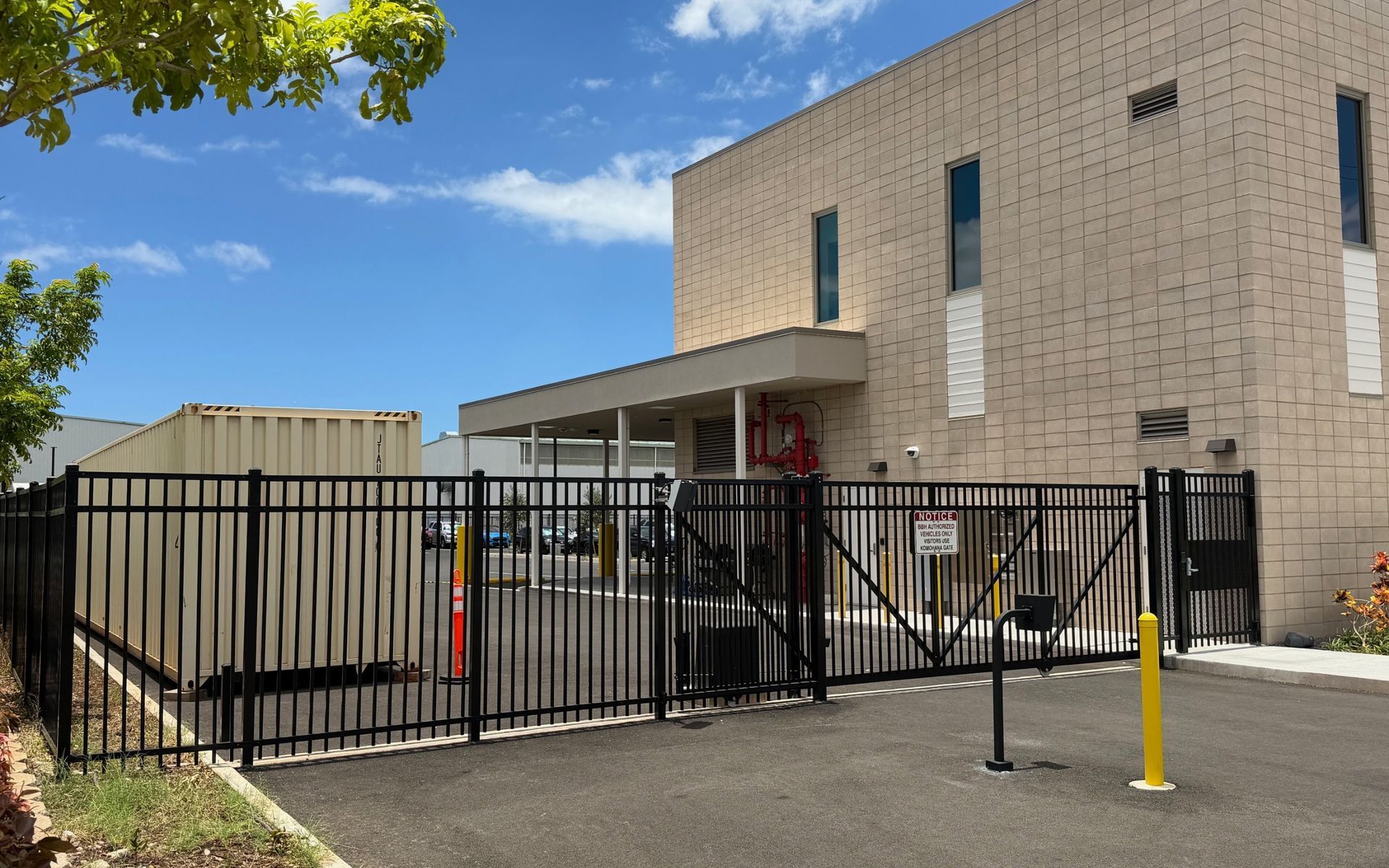 Gated entrance to a beige building under a blue sky, with a small tree and yellow bollard in front