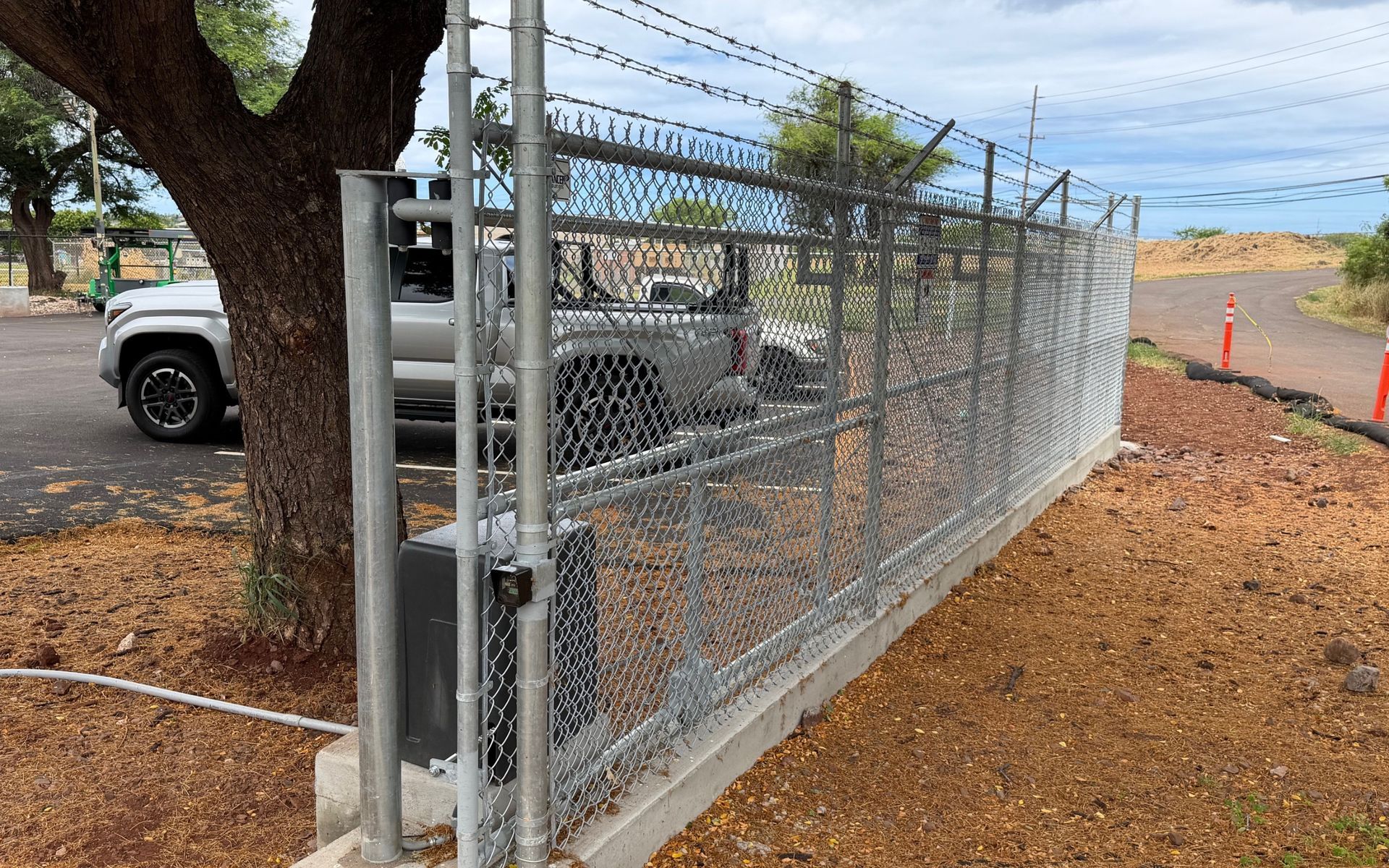 Chain-link fence beside a tree and parked truck, with barbed wire along a gravel roadside.