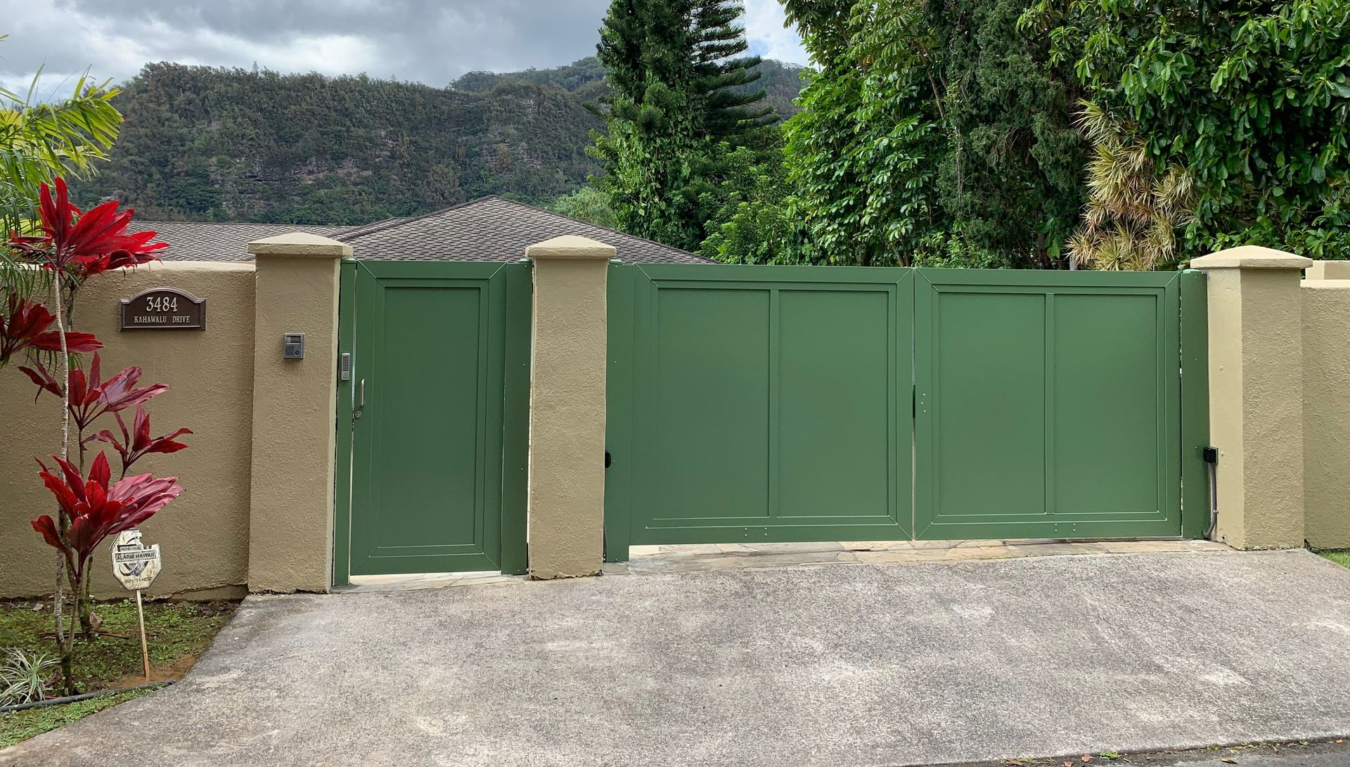 Green gated entrance with tan columns and a driveway, lush green foliage and mountains in the background.