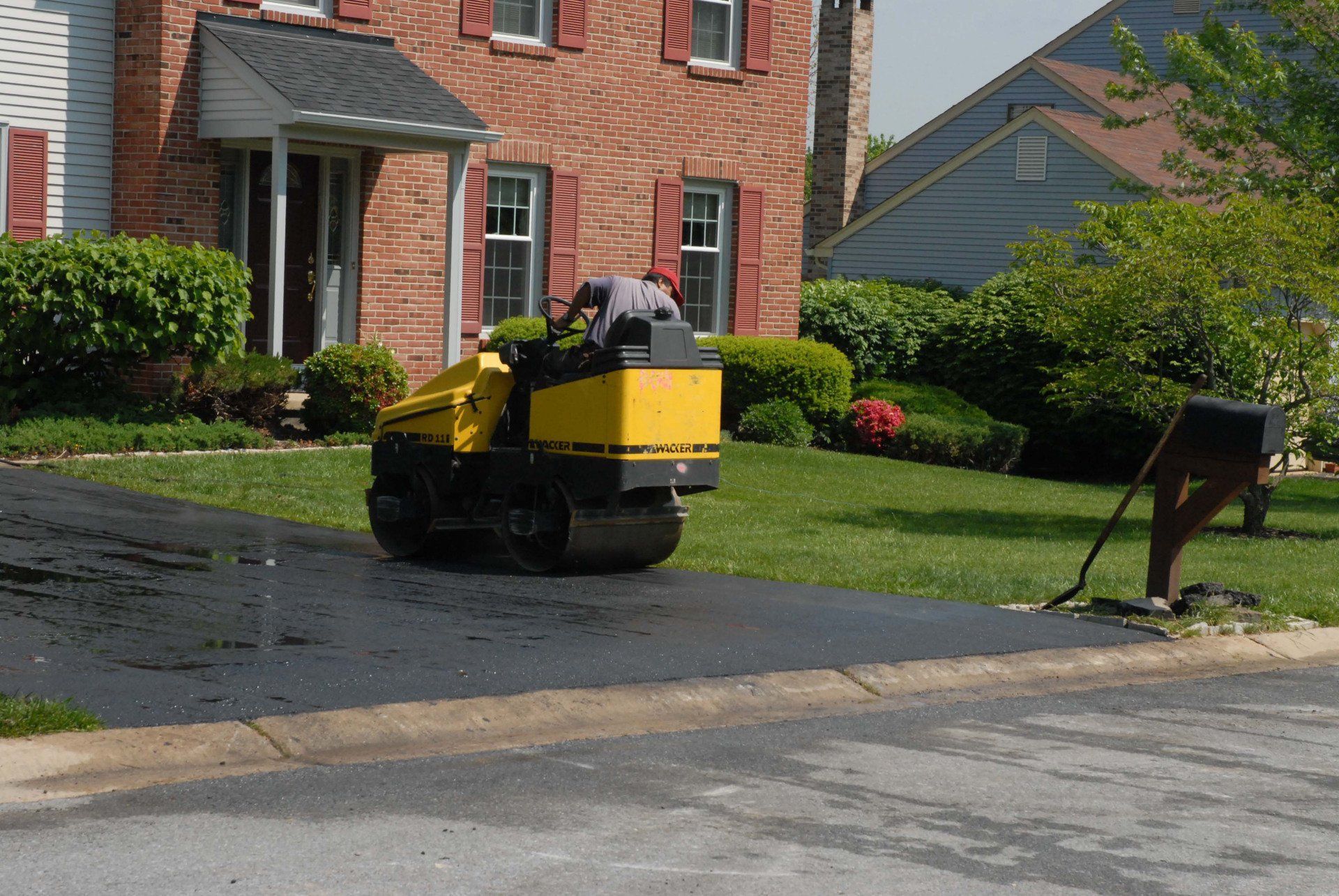 A yellow roller is being used to asphalt a driveway in front of a brick house.