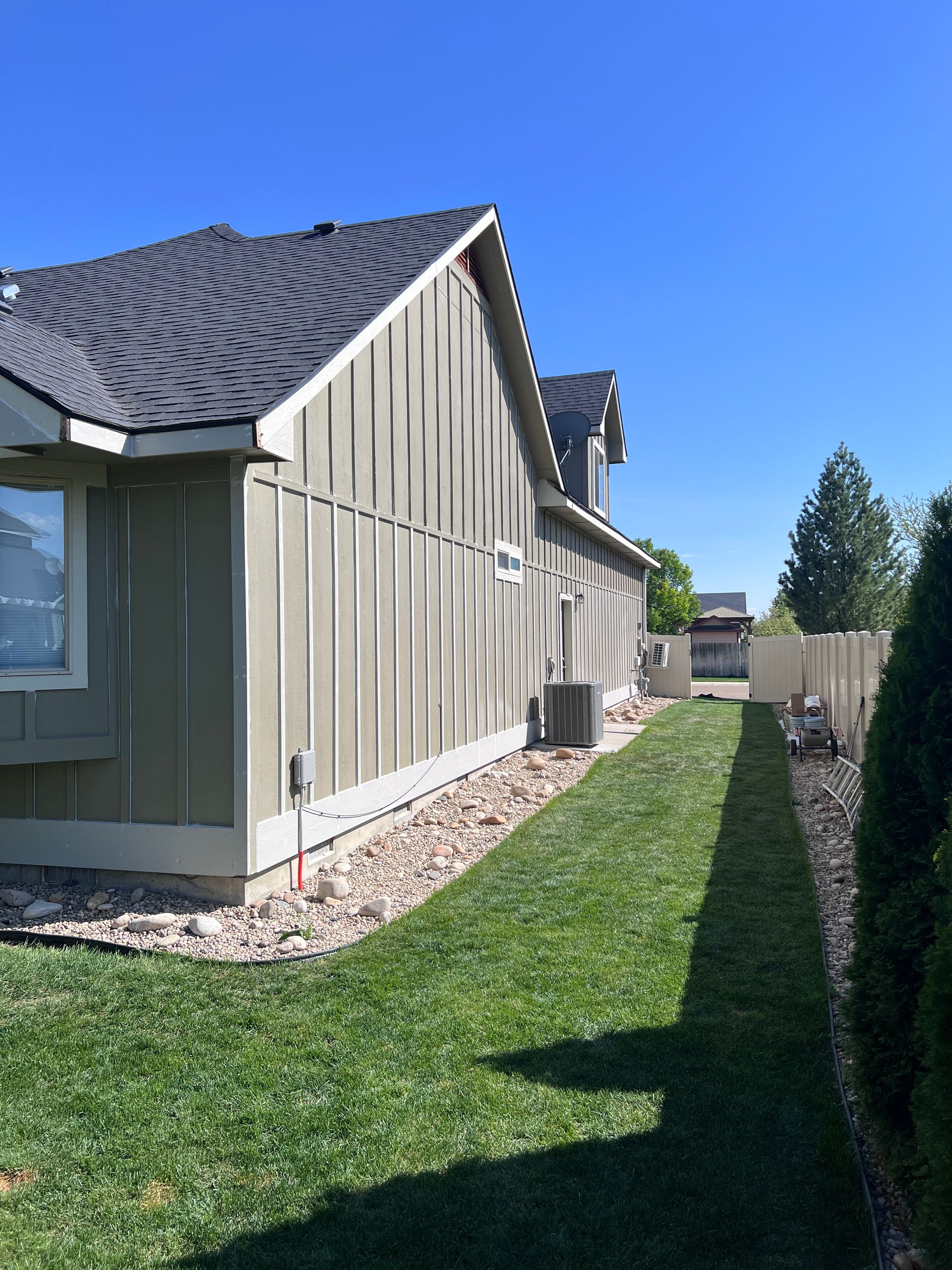 Side of a beige house with a dark roof, green lawn, and a narrow walkway beside a wooden fence.