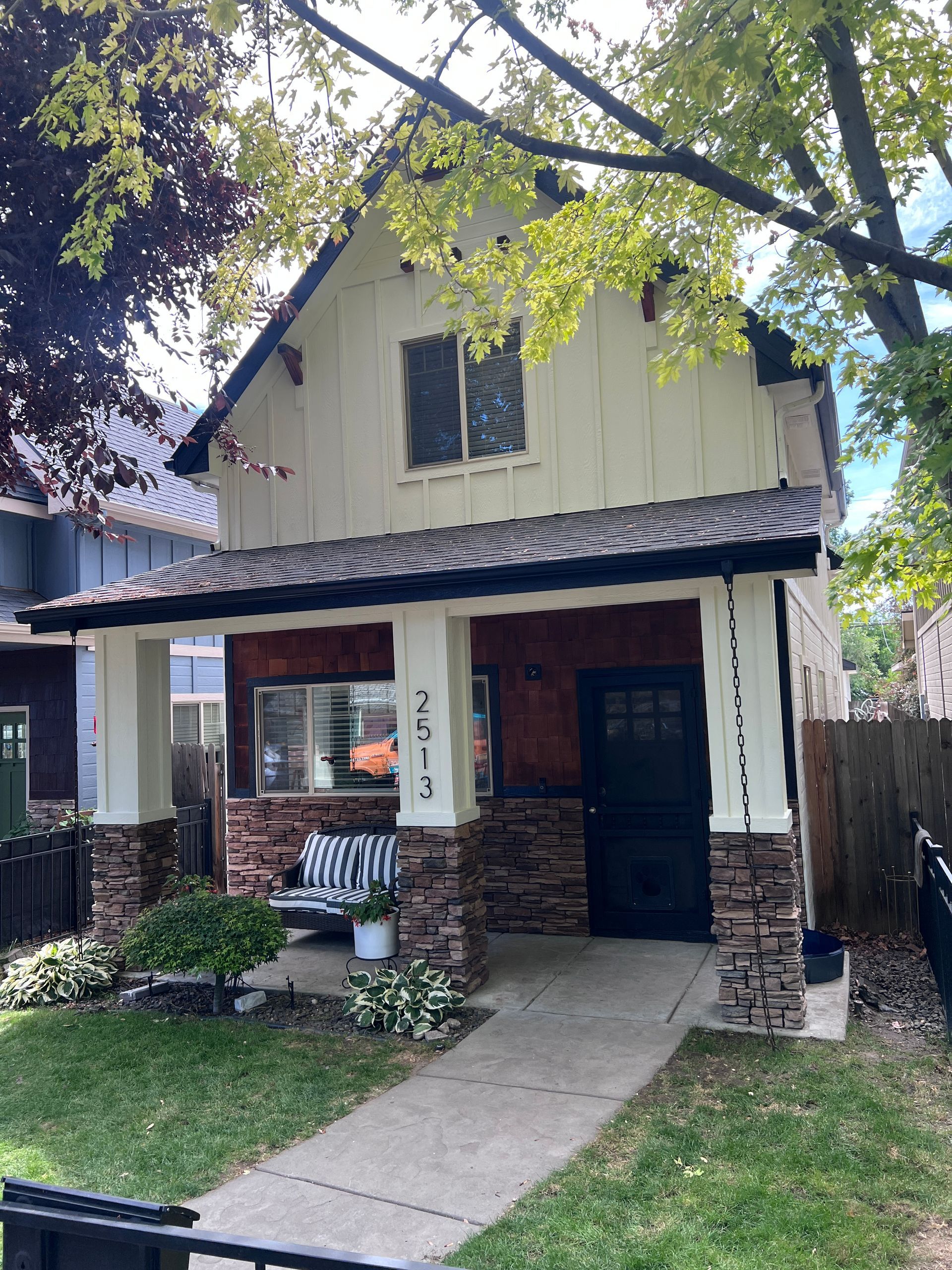 Two-story house with a front porch, brick columns, and a walkway under leafy trees.
