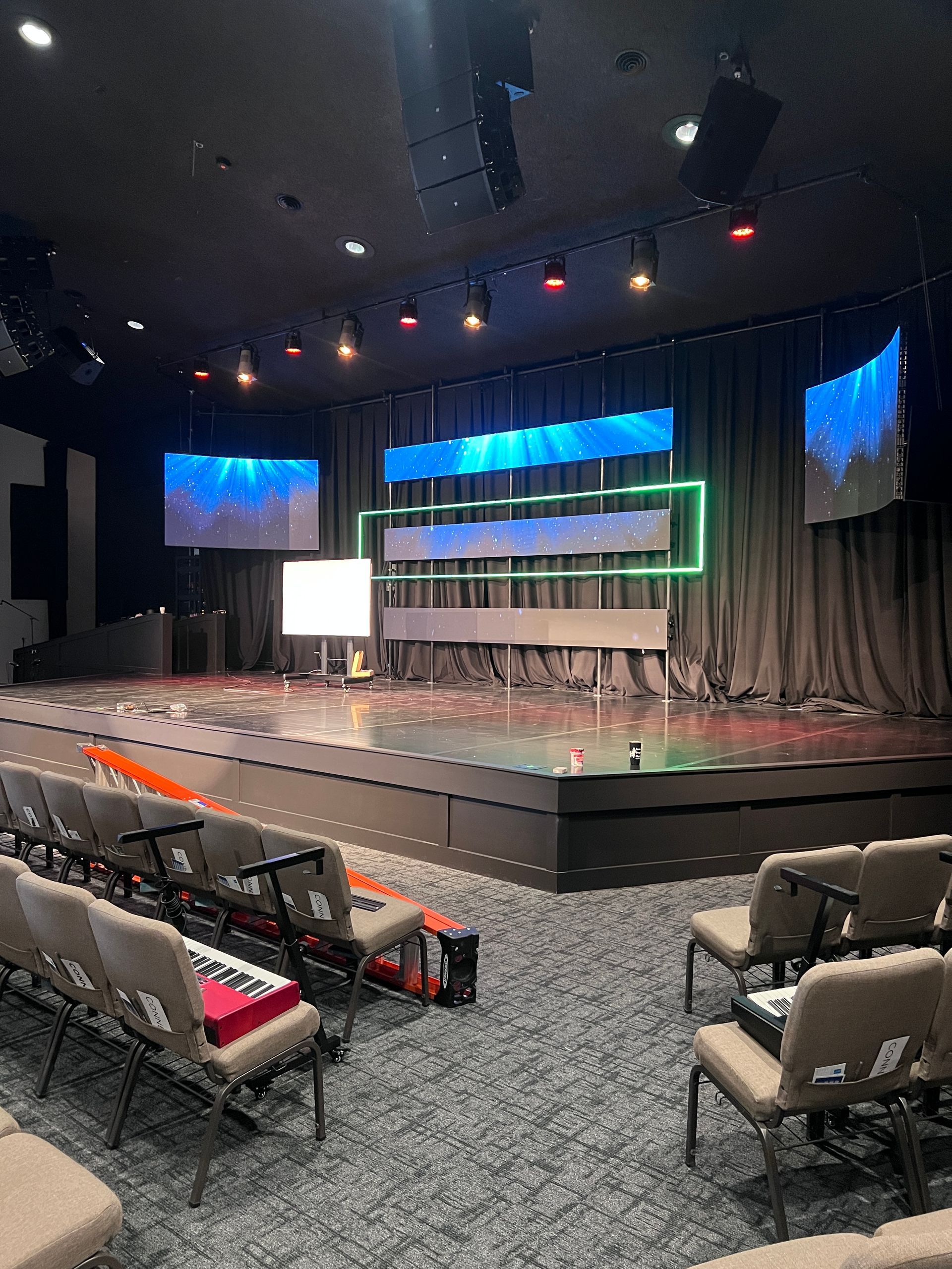 Empty theater stage with blue and green lighting, rows of chairs, and a carpeted audience area