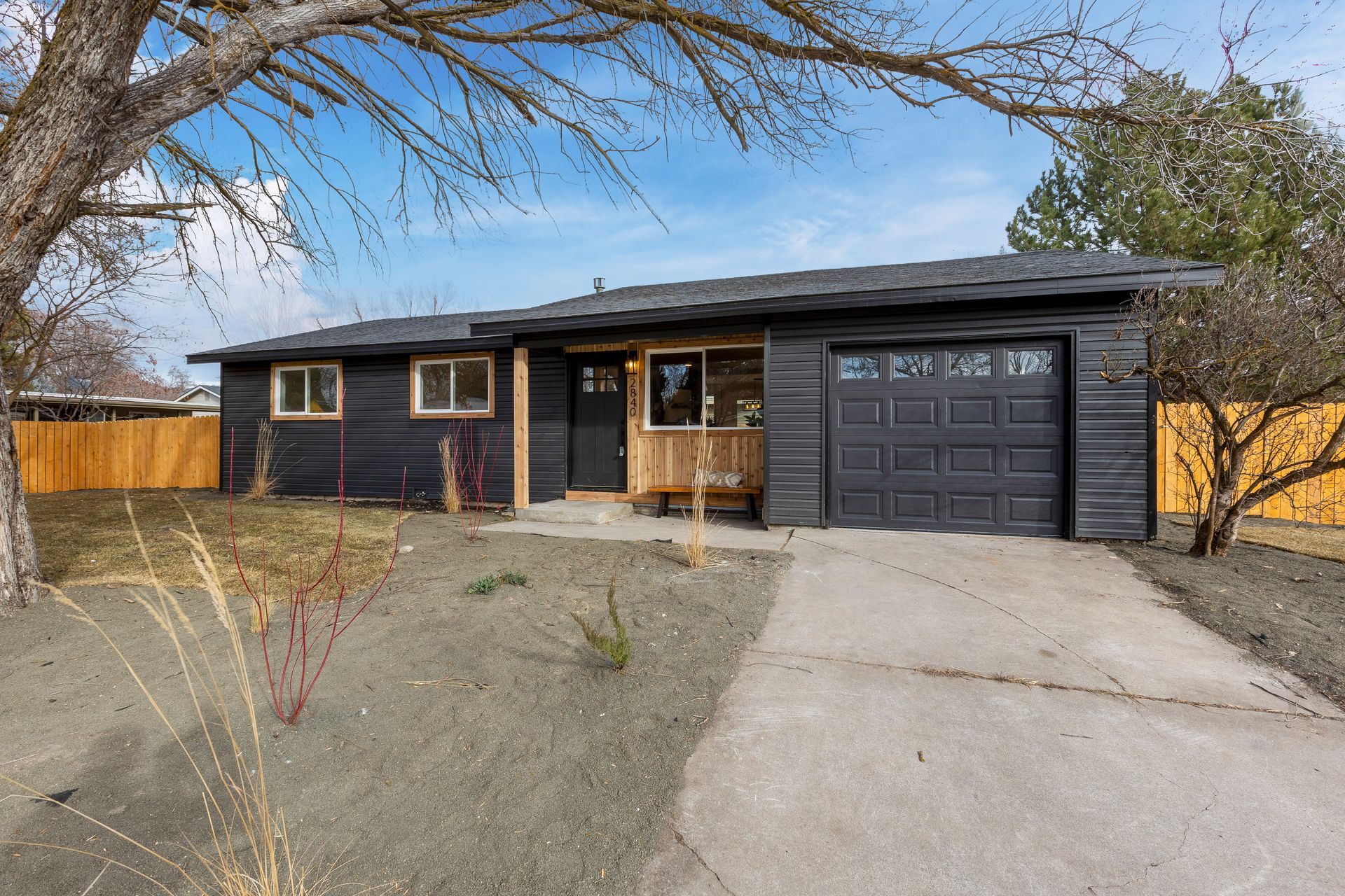 Single-story house with a black garage door, front porch, and concrete driveway under bare trees.