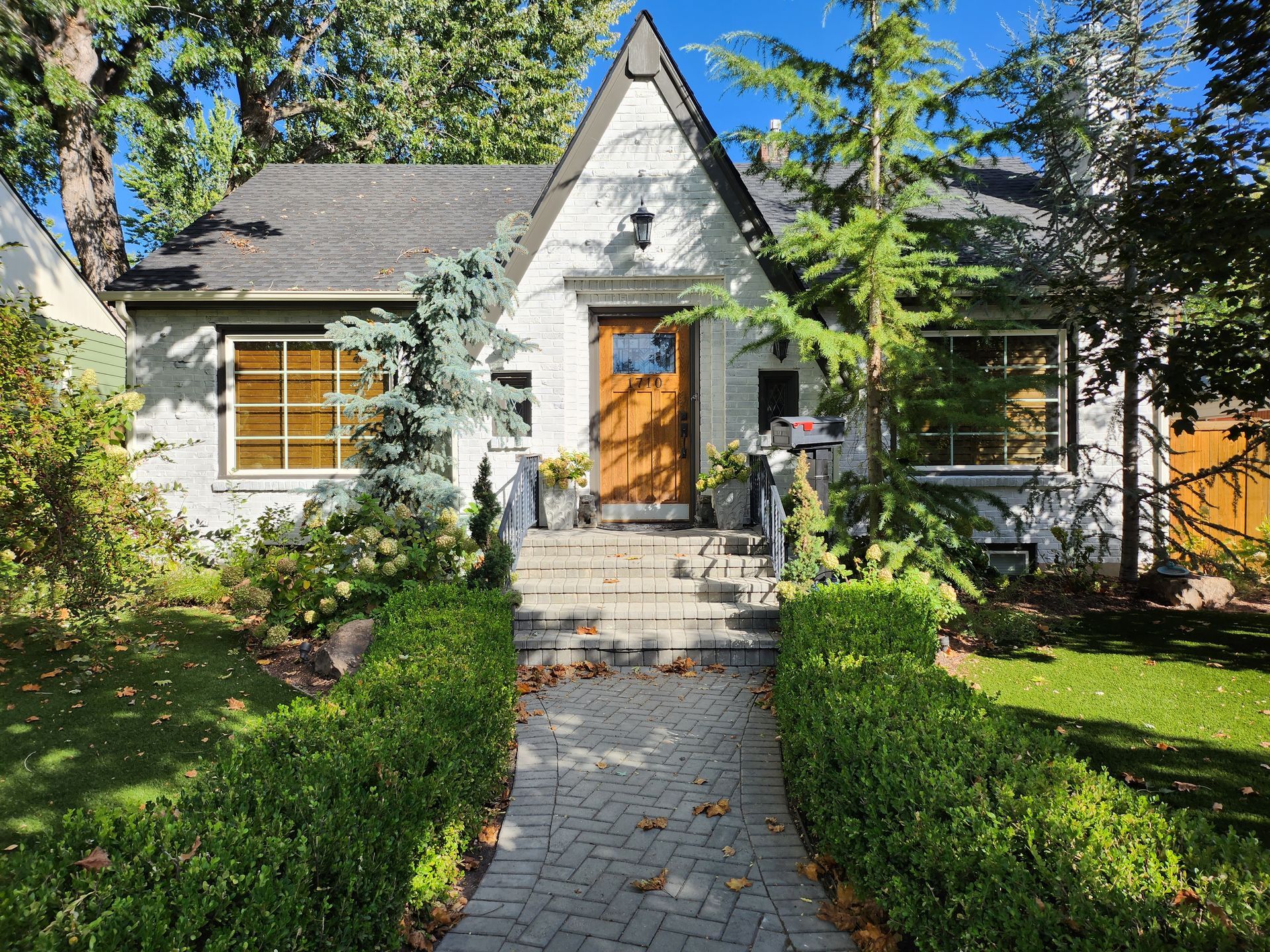 Small cottage with steep roof, front steps, and a brick path amid green hedges and trees