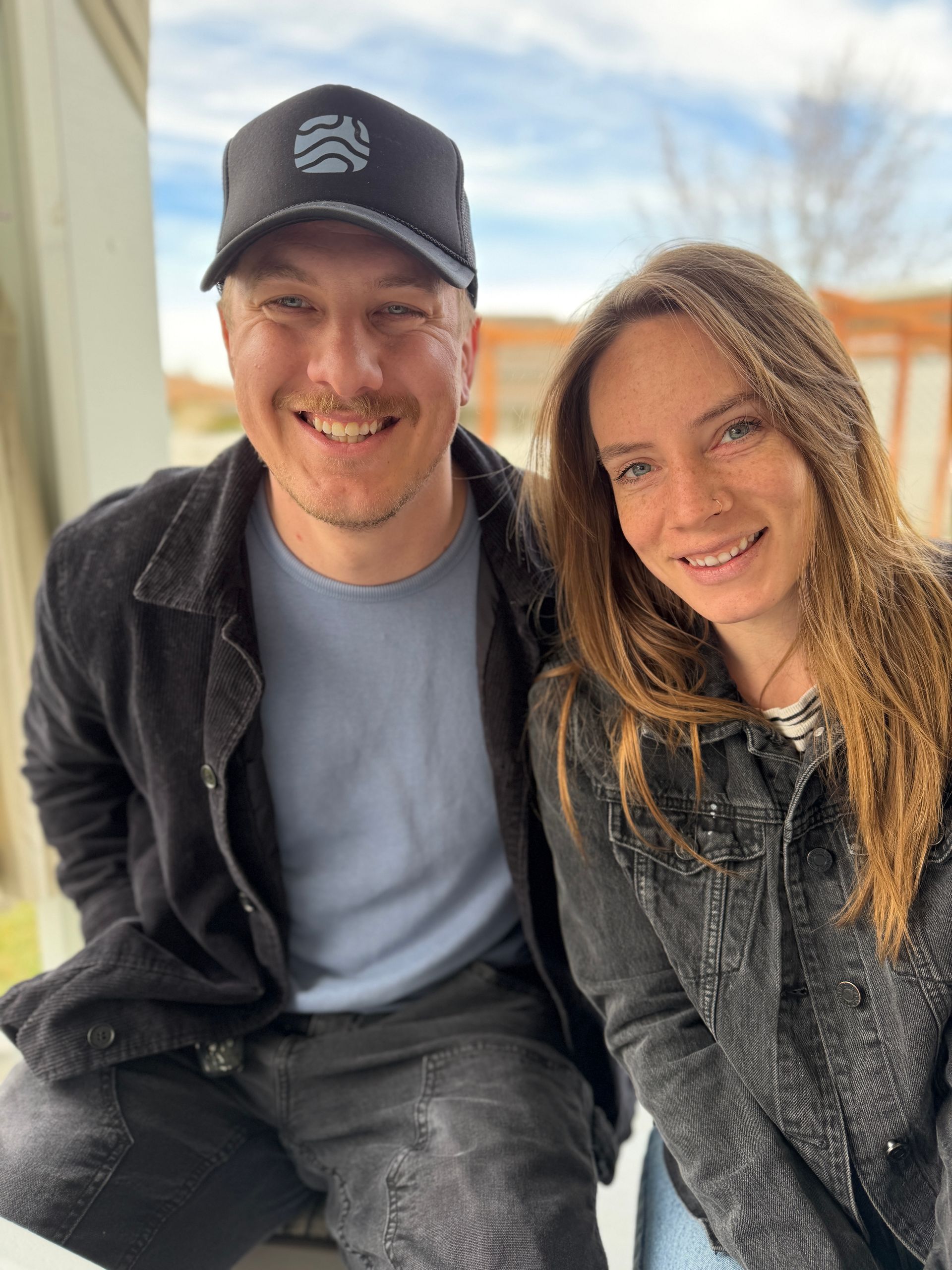Two smiling people sitting close together indoors, wearing casual dark jackets and a cap.