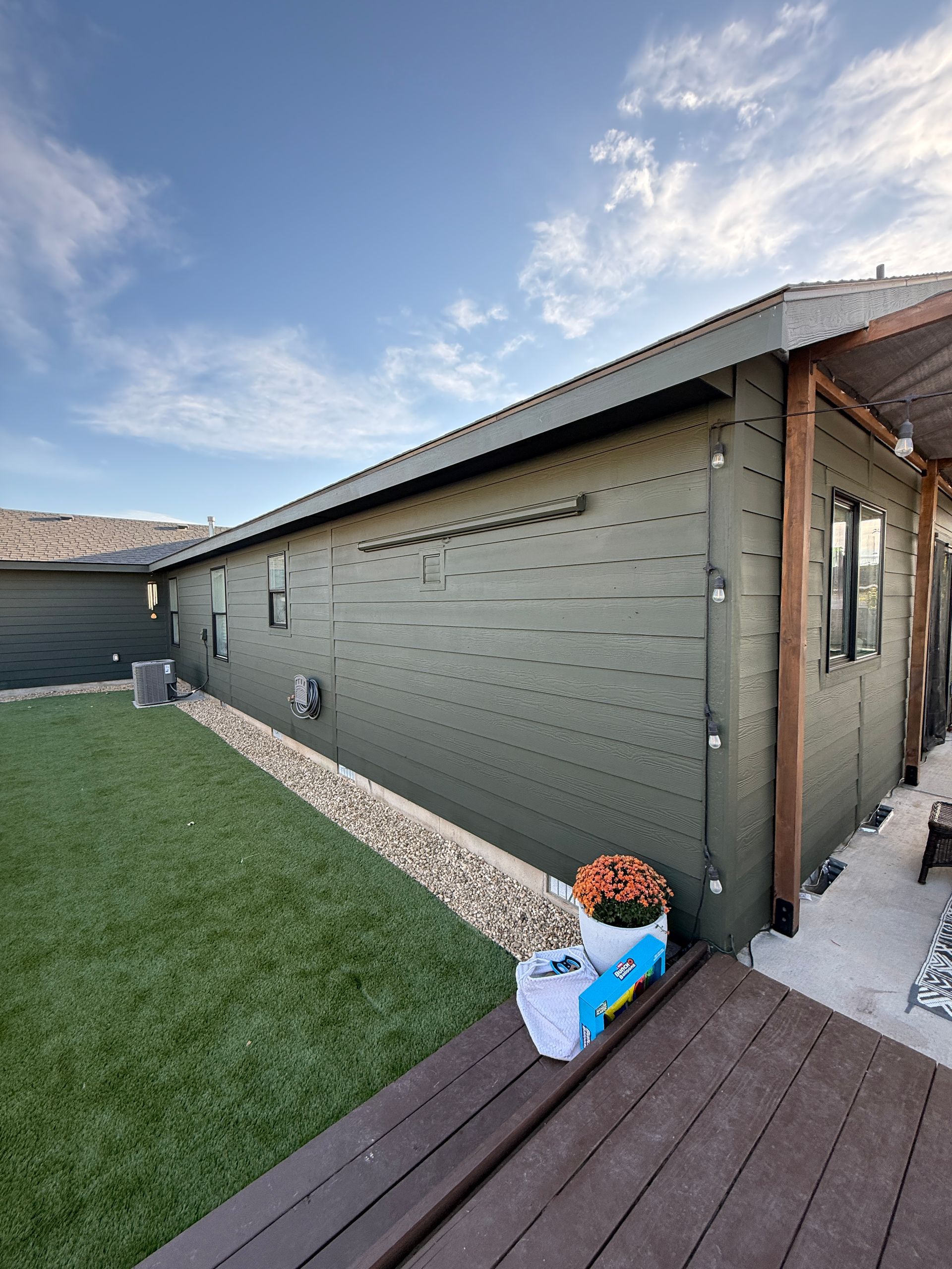 Backyard view of a beige house wall, green lawn, wooden deck, and potted flowers under a partly cloudy sky.