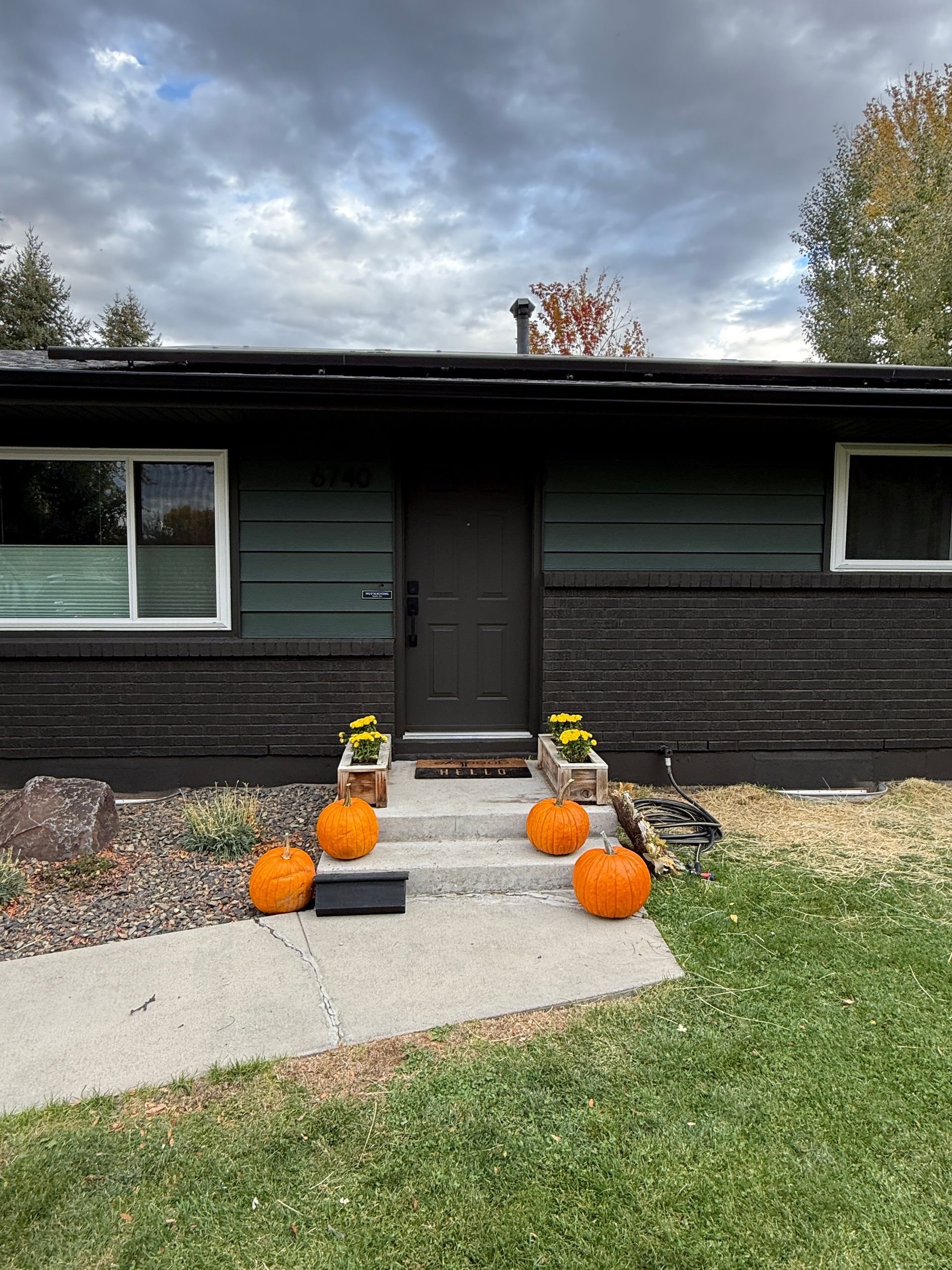 Front of a dark house with pumpkins and mums on a gravel walkway under cloudy skies